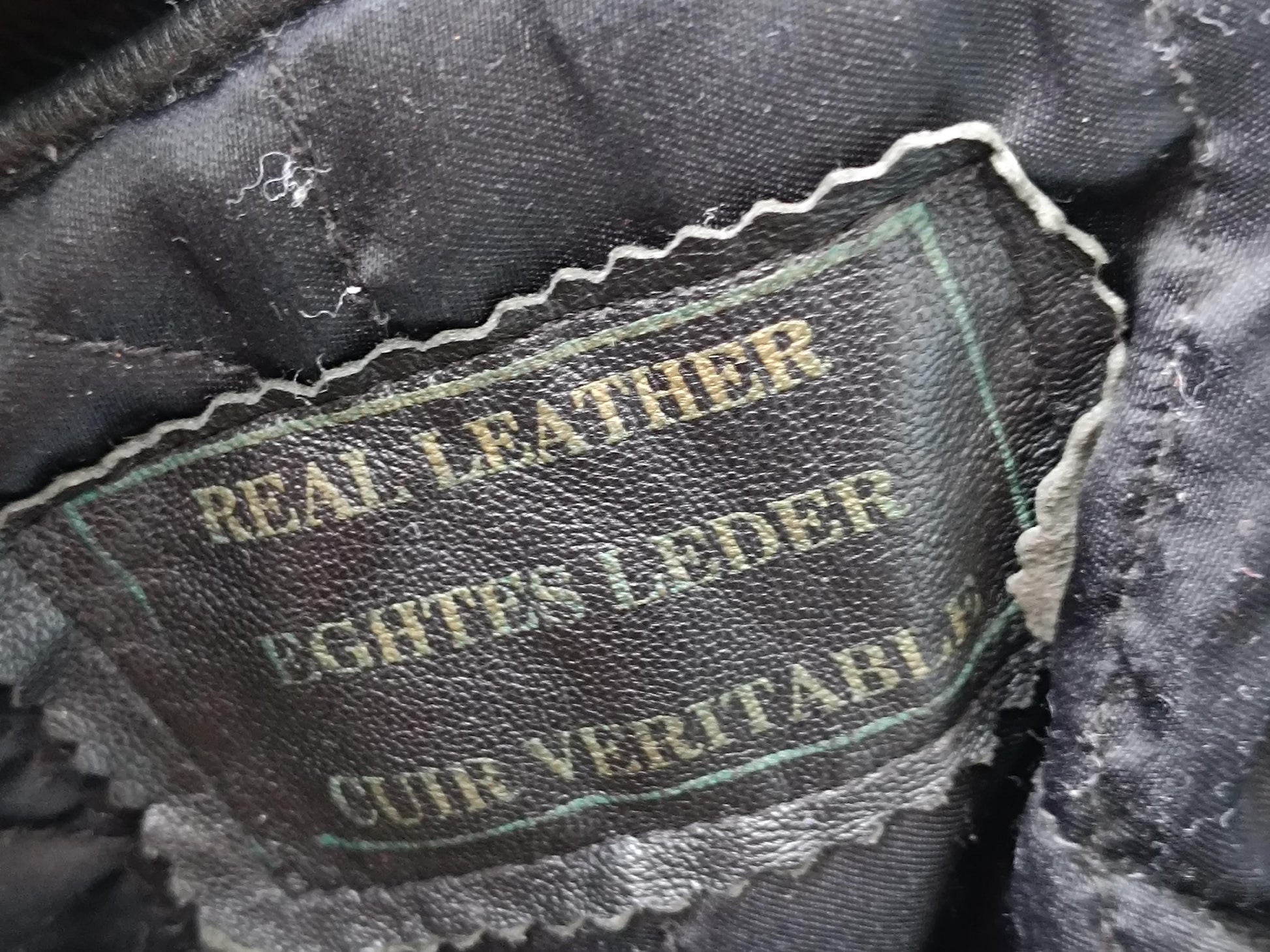 A photograph of a  black fabric label of a rock and roll style vintage leather jacket. The label reads Real leather in English, German and french.