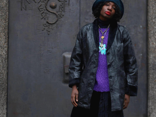 Female model wearing a 90s vintage black patchwork leather coat, purple textured sweater, and a dark skirt, with layered necklaces and a dark hat. She is photographed against a textured gray wall. The faux-fur leather jacket is by the brand Anne De Lancay and gives off a boho rock n roll  vibe.