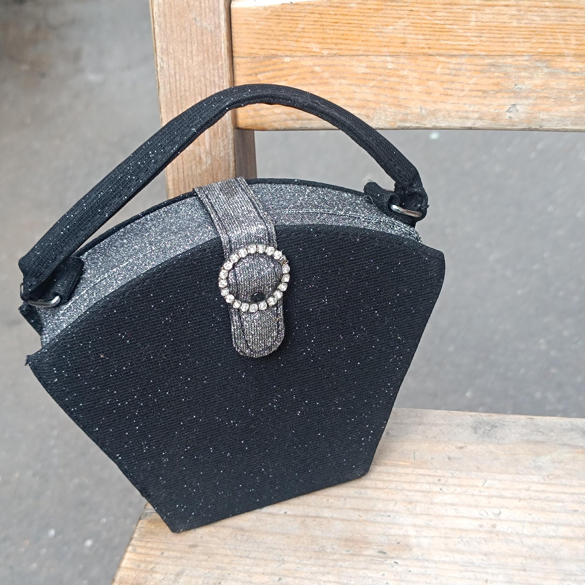 Y2K-inspired black mini bag with a glittery surface, a top handle, and a faux-bling circular detail on the closure. The bag&#39;s unique pentagon shape is visible against a wooden background.