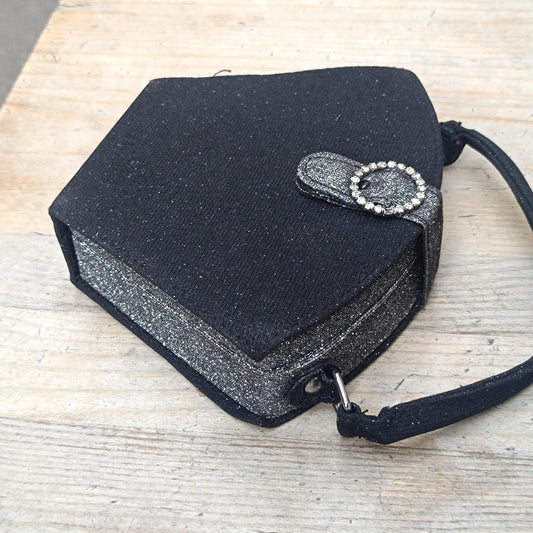A black, pentagon-shaped mini bag with a shimmering glitter finish. It features a top handle and a tab closure with a circular faux-bling embellishment. The bag is photographed from a top-down perspective on a rustic wooden surface.