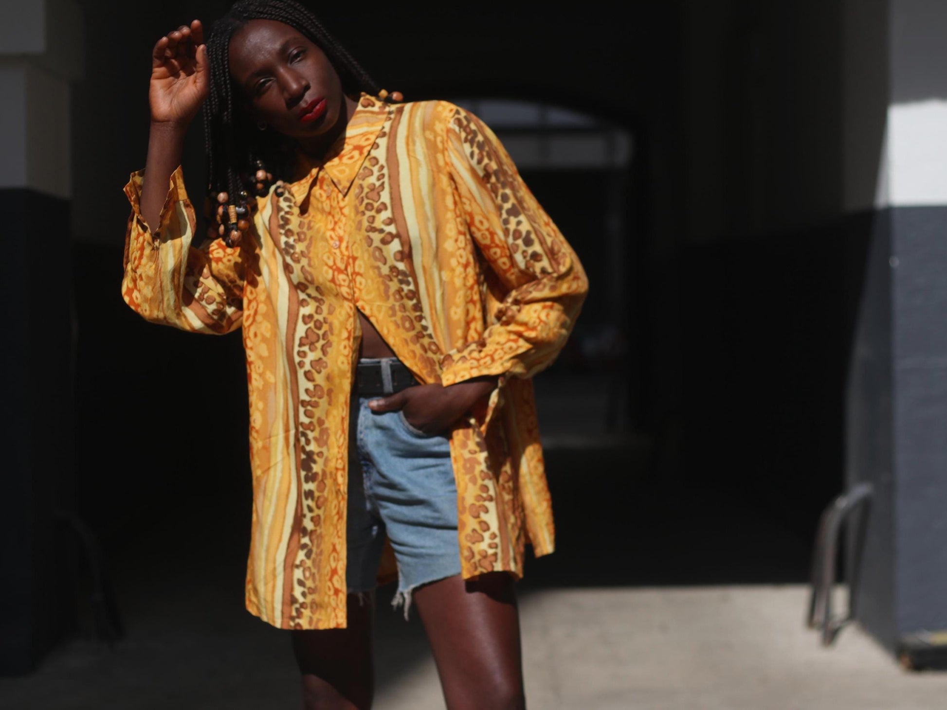 A woman with braided hair with beads modeling a vibrant yellow vintage blouse. The 90s blouse features a bold animal print with swirling patterns in shades of yellow, gold, brown, and cream. It has long sleeves, a button-down front, and a loose, oversized fit. She pairs it with light blue denim shorts. She lok=oks directly into the camera with one hand raised.