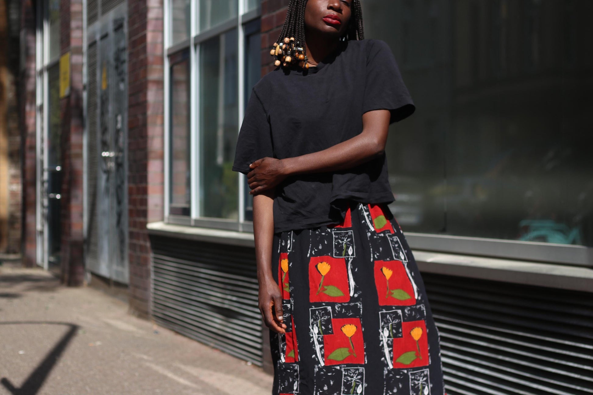 A female model in a calm pose with on arm  touching the elbow of the other, wearing a striking 90s red floral vintage maxi skirt, surrounded by urban architecture and soft afternoon sunlight.