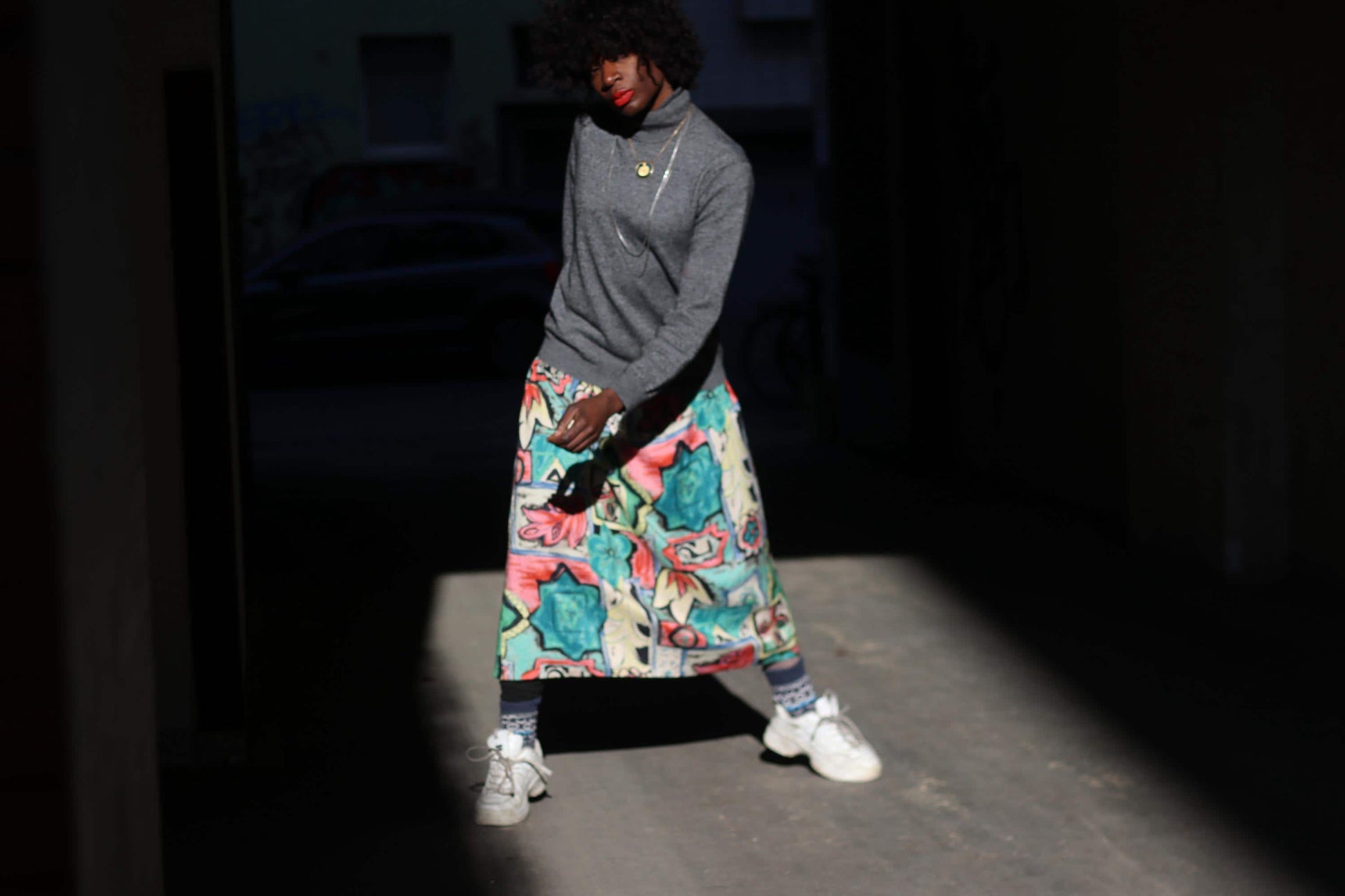 A female model with curly black hair wears a mid length  multicoloured vintage skirt with a unique, artistic print against a dark, shadowy background. They are dressed in a gray sweater and white sneakers