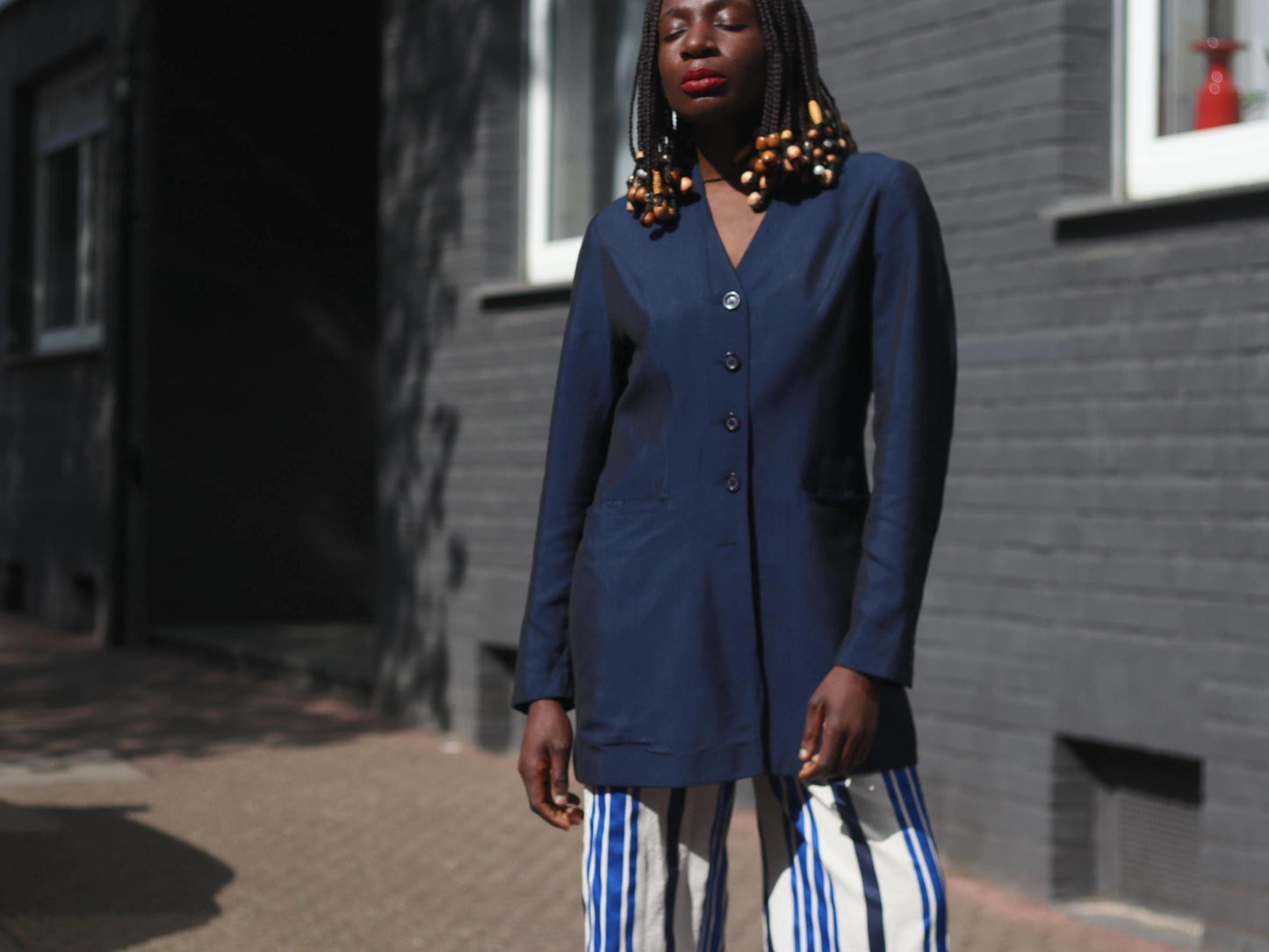 A female model with beaded braids is photographed on a sunny day against a brick wall on a sunny day.in a vintage 90s blue blazer by the brand Bonita styled with blue and white striped cullotes . The look is a vintage twist on power dressing. s