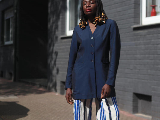 A female model with beaded braids is photographed on a sunny day against a brick wall on a sunny day.in a vintage 90s blue blazer by the brand Bonita styled with blue and white striped cullotes . The look is a vintage twist on power dressing. s