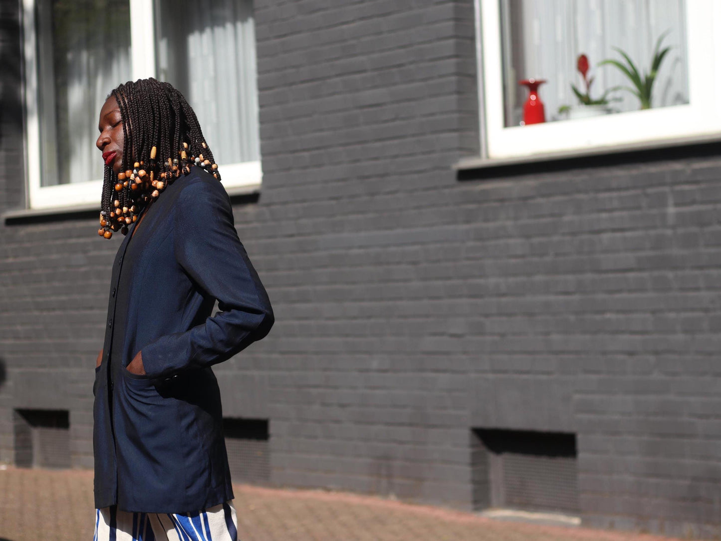 A side view of a female model with beaded braids in a vintage retro blue blazer and striped pants standing on a sidewalk with a dark brick wall behind her.