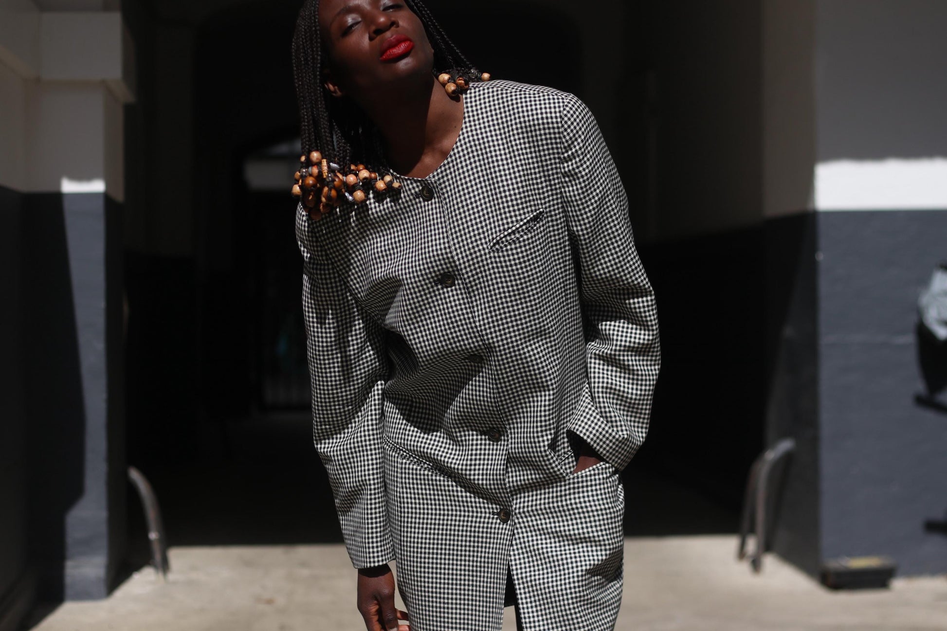 Female Model with beaded braids wearing a vintage checkered  90s blazer as a dress, photographed outdoors on a sunny day in an alley with dramatic lighting
