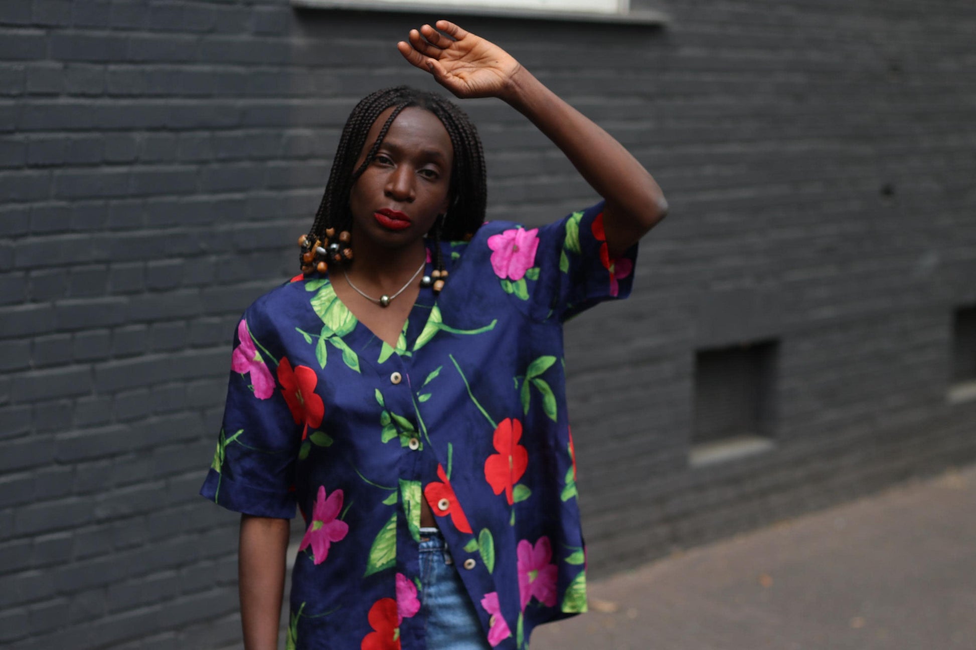 An editorial-style  image of a model in a vintage navy floral blouse with bold hibiscus print photographed Infront of a grey brick wall with one hand up in the air with a melancholic expression