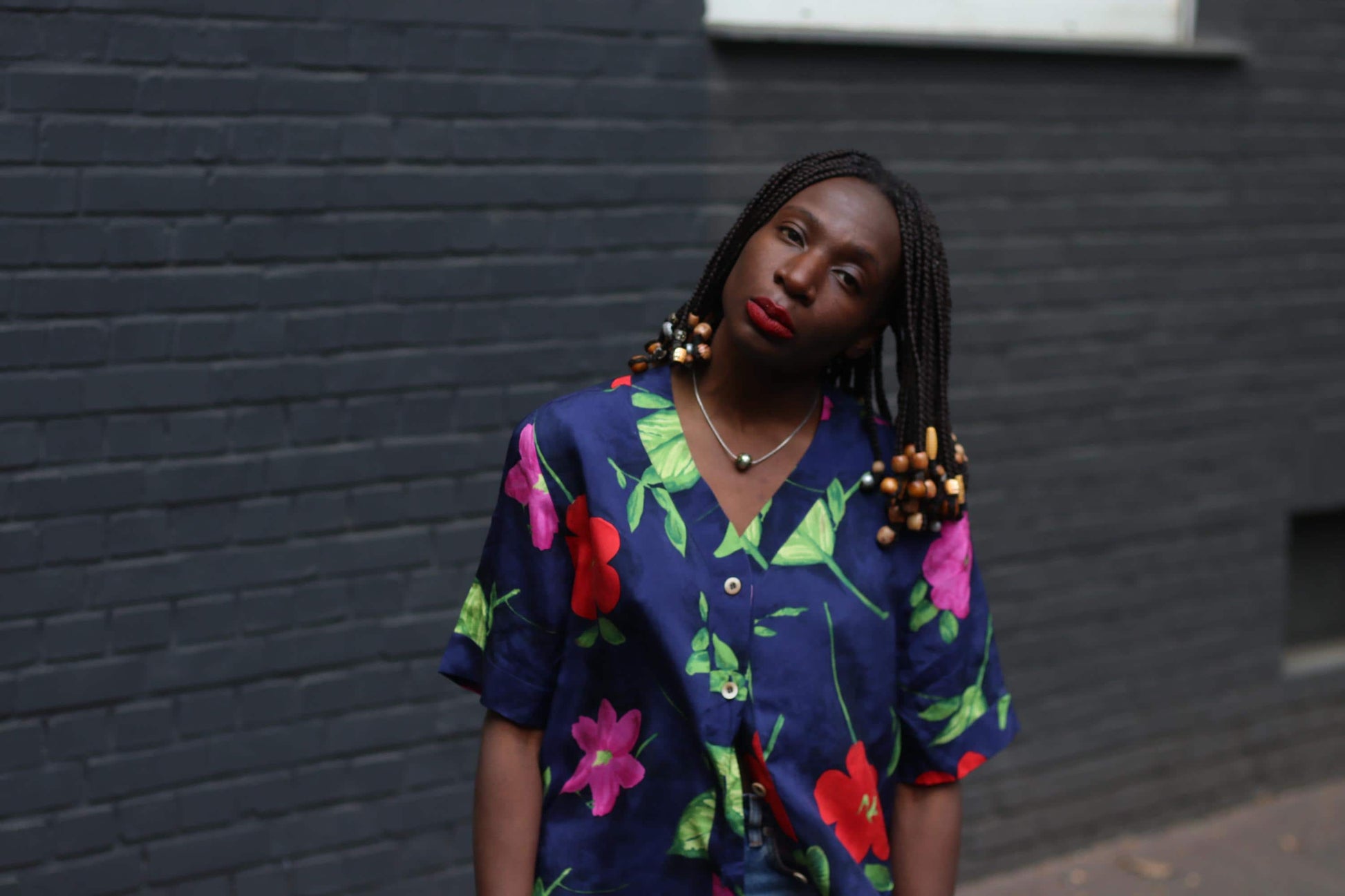 Front view of model of African heritage with beaded braids in a vintage navy floral blouse with bold hibiscus print photograohed outdoors against a grey brick wall