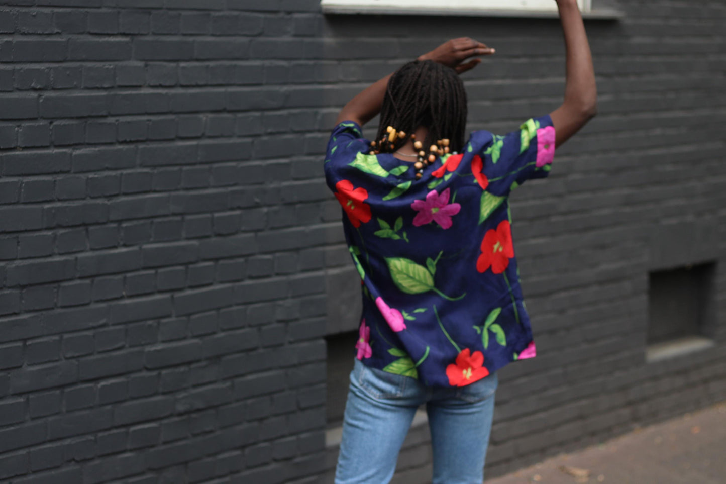 Back view of vintage navy floral blouse with bold hibiscus print showcased on a model. of African heritage with beaded braided hair. The blouse is styled with blue jeans and model has both hands up. in the air.