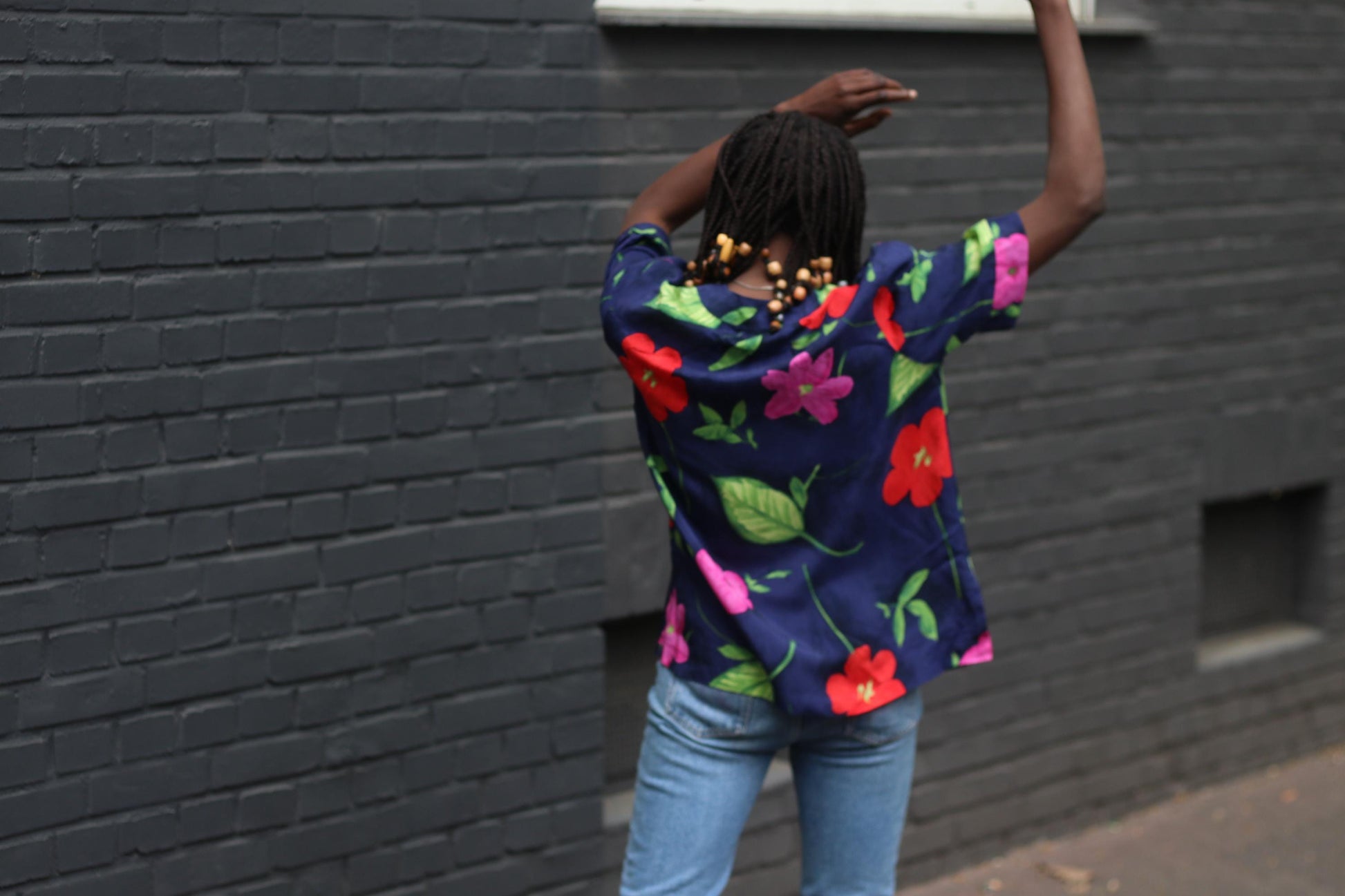 Back view of vintage navy floral blouse with bold hibiscus print showcased on a model. of African heritage with beaded braided hair. The blouse is styled with blue jeans and model has both hands up. in the air.