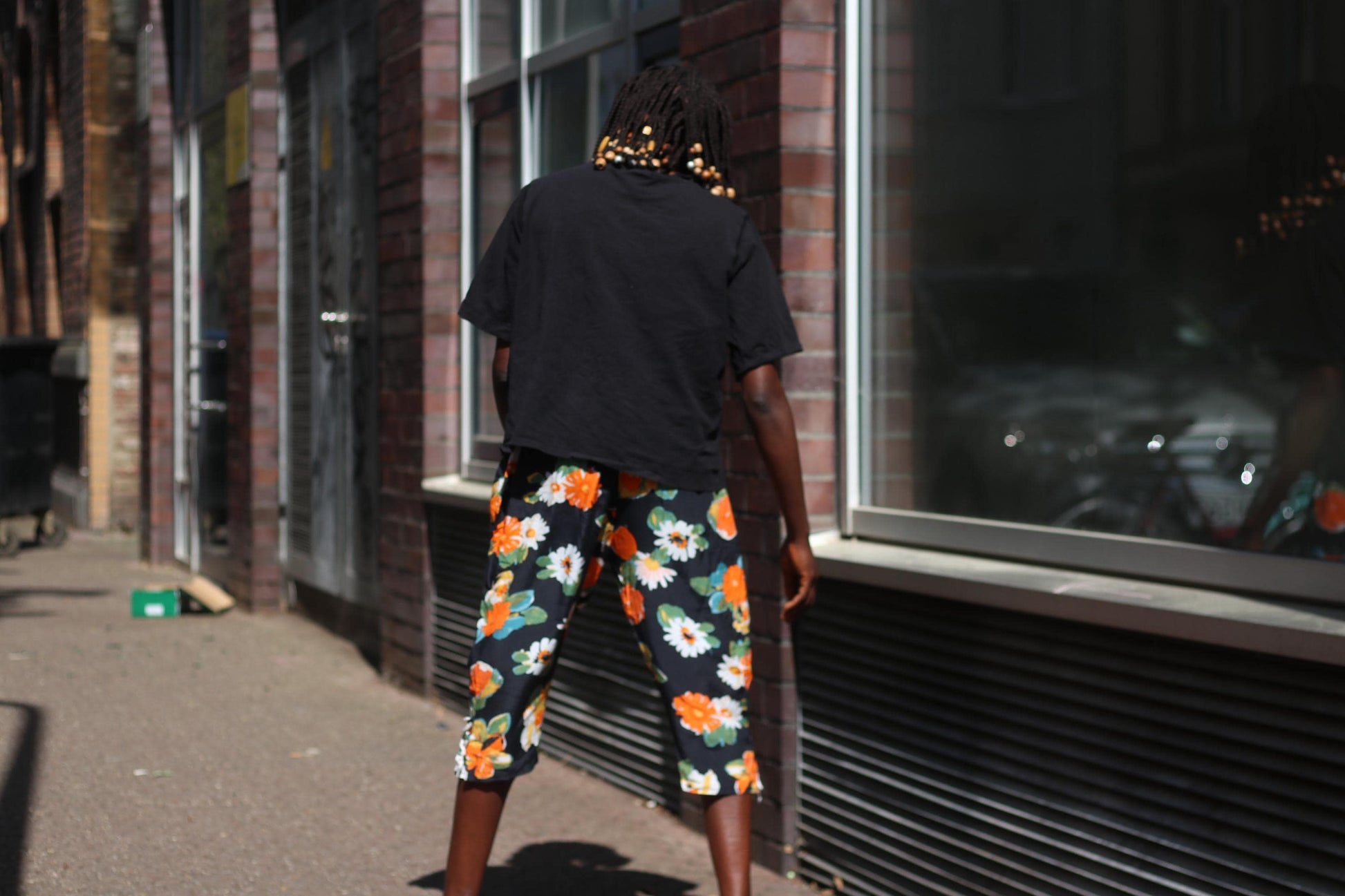 A back view of a female model standing in bright sunlight wearing vintage black capris from the 90s with bold orange and white floral prints, paired with a simple black t-shirt  against a brick wall and glass storefront.