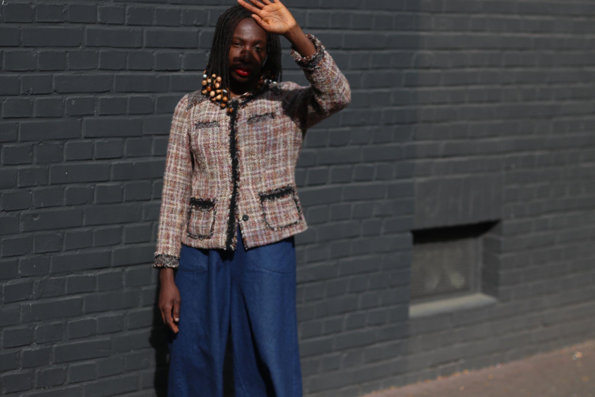 Female model in a vintage textured 90s tweed blazer with wide blue pants, standing against a solid dark grey brick background with one hand up as I'd to shield face from the sun