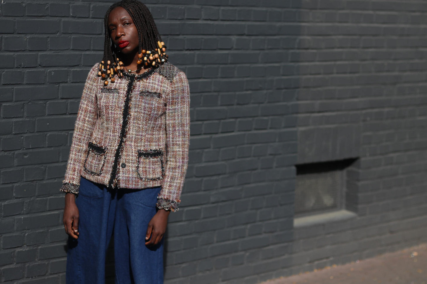 Front view perspective of a fashionable look featuring a tweed blazer in shades of brown, cream, and black worn with loose-fitting blue trousers in an outdoor setting with a dark brick wall