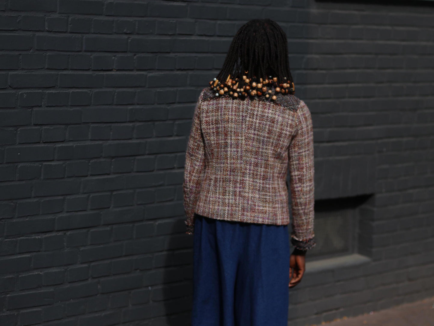 Back view perspeview of  a model in a vintage 90s tweed blazer in shades of brown, cream, and black with loose-fitting blue jeans in an outdoor setting with a dark brick wall