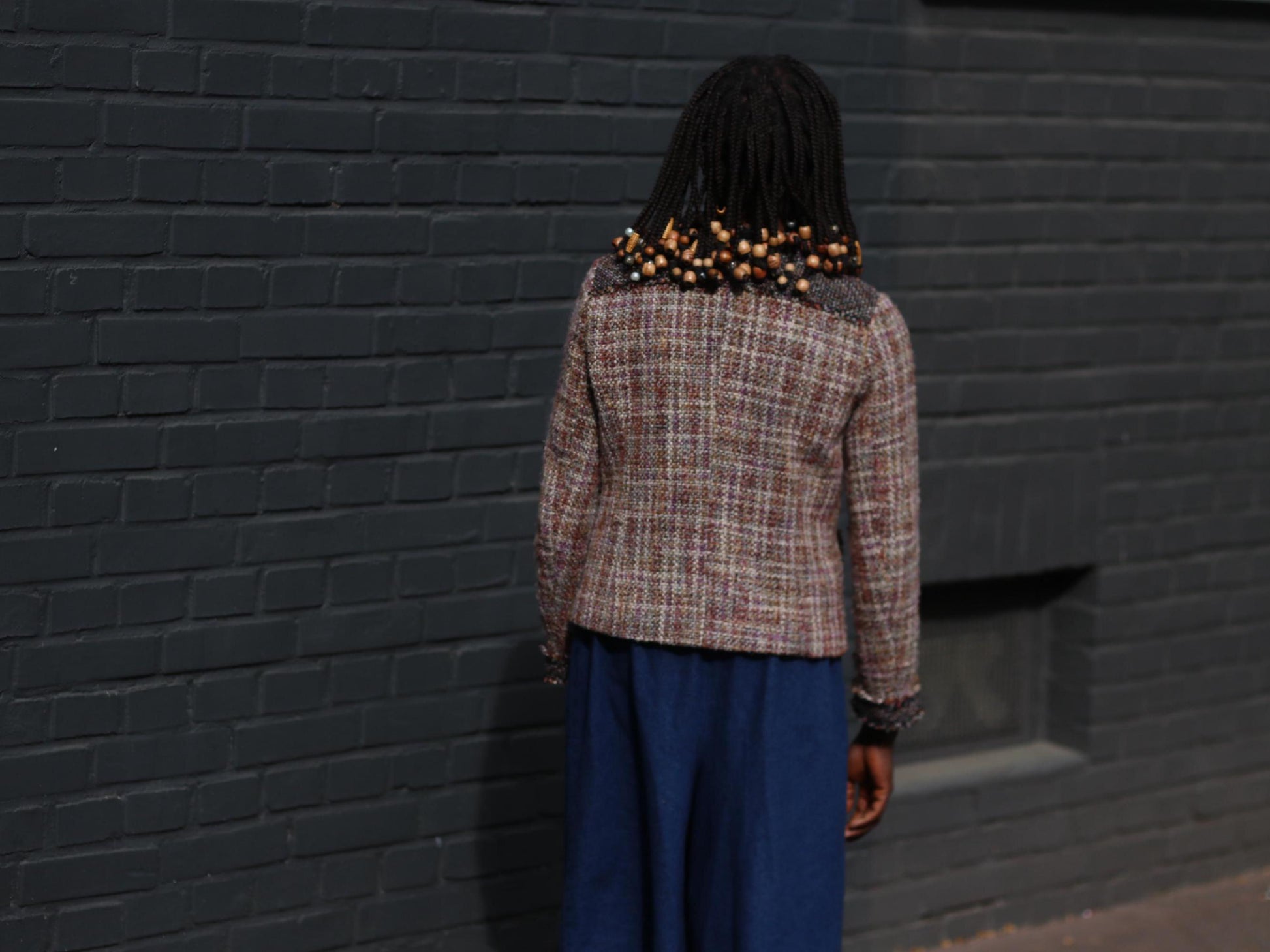 Back view perspeview of  a model in a vintage 90s tweed blazer in shades of brown, cream, and black with loose-fitting blue jeans in an outdoor setting with a dark brick wall