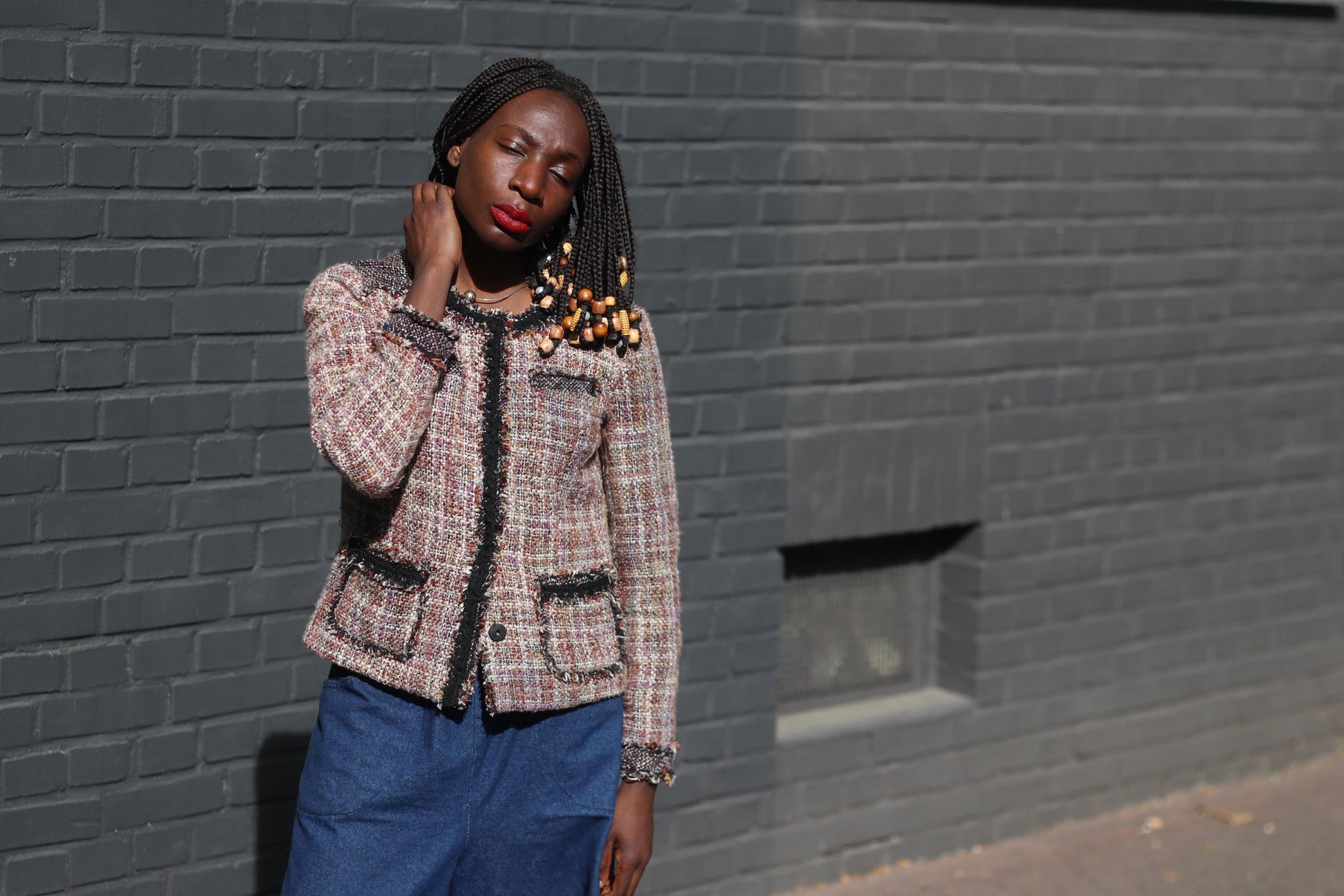 Female model of color with beaded braids   in a brown textured tweed blazer from the brand Coldwater Creek styled  with wide blue pants, standing against a solid dark grey brick background.