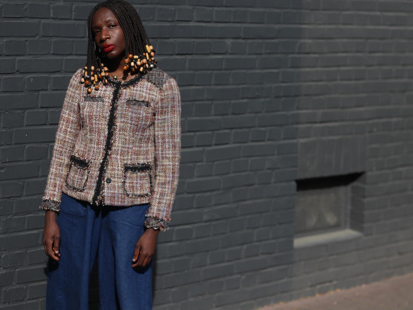 Model showcasing a brown, cream, and black tweed blazer featuring a round neckline, black edging, and gold accents, complemented by flowing blue trousers in an urban setting.
