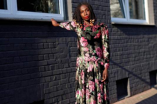 A model of African heritage with braids with beaded details in  a vintage 90s  floral velvet midi dress  with a drop waist pleated silhouette leans  her hands against  a black  bricked wall