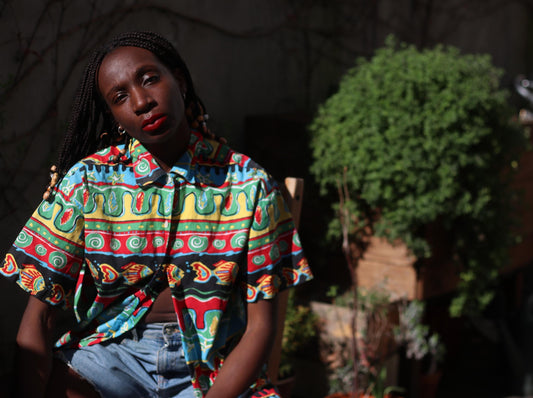 A female model with long, dark braids with beads is seated outdoors, wearing a vintage 90s short-sleeved shirt with a vibrant, multicolored pattern. The shirt features red, yellow, green, and blue hues. They are also wearing light blue denim jeans. The background shows a blurred outdoor setting with plants and a wooden structure.