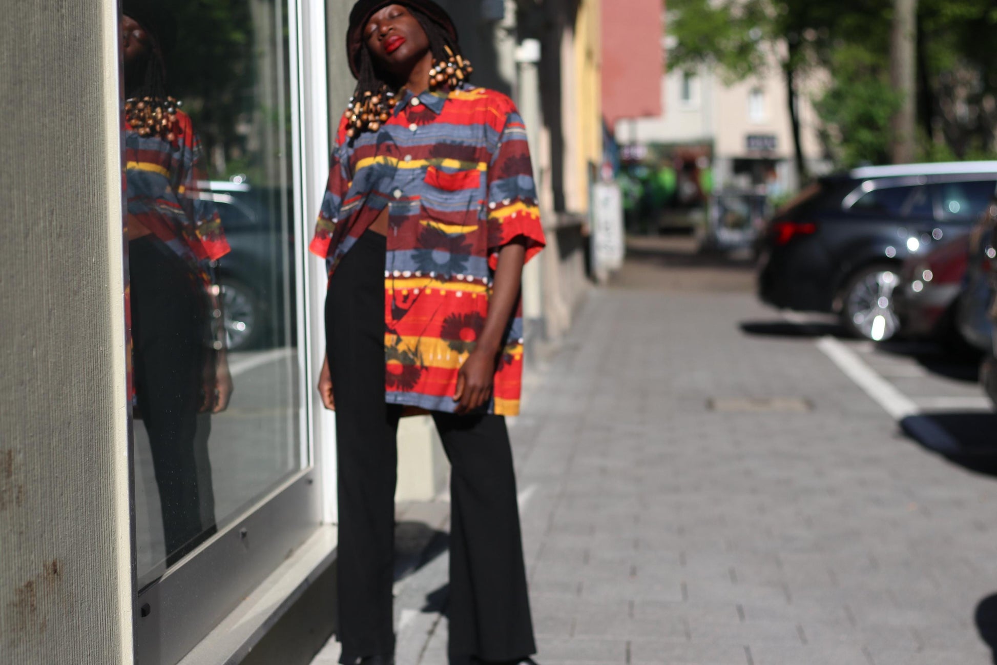 Artistic photograph of a female model with beaded braids in a 90s vintage Montino tropical floral shirt with a retro bold island print, black pants and hat captured in an urban street setting with cars and buildings in the background. The image highlights the shirt&#39;s red and yellow hues, short sleeves, unisex fit, and bold summer tones.