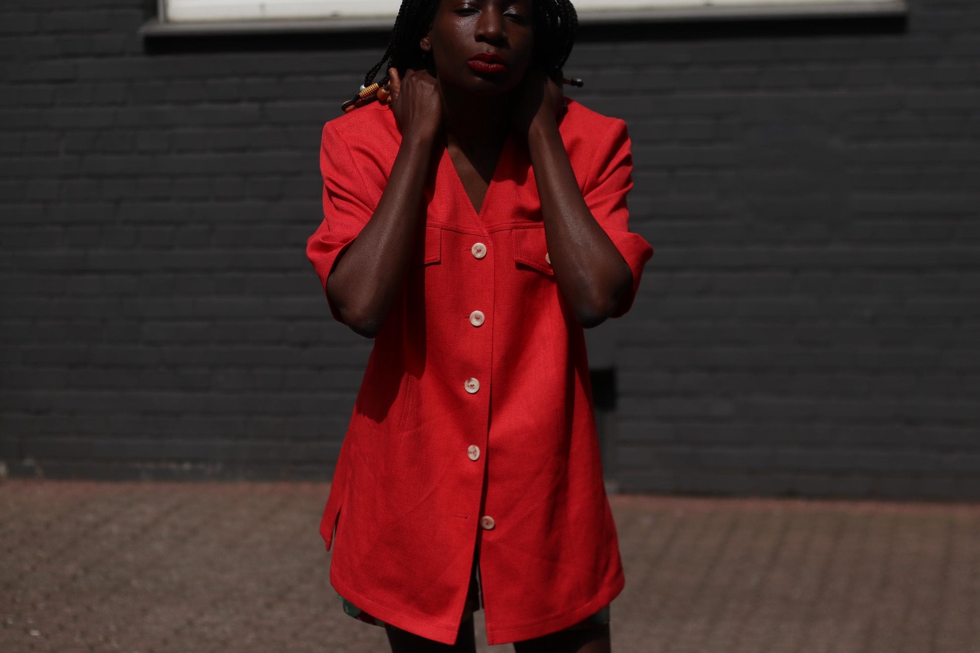 Female model in a vintage 90s Short-Sleeve Blazer by Mark Wald as a short dress photographed in front of a grey brick wall.  The image showcases the standout features , from the v-Neck silhouette to the buttons and chest pockers
