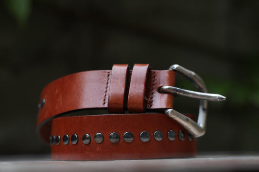 A vintage brown leather belt with rows of metal studs displayed on a textured surface with greenery in the background