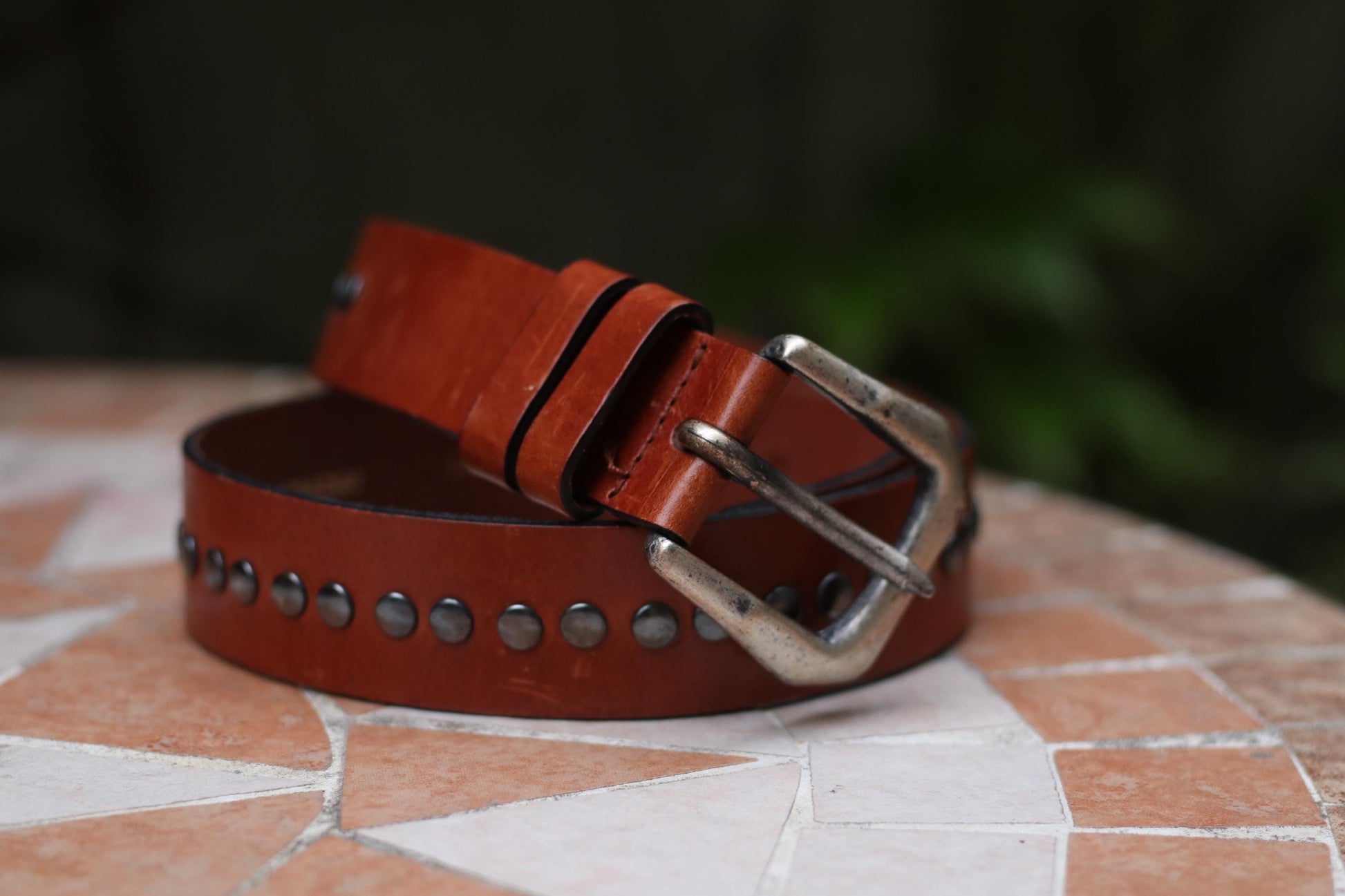 An artistic shot of a studded brown leather belt from the 70s on a tiled table outdoors with greenery in the background creating a rustic setting