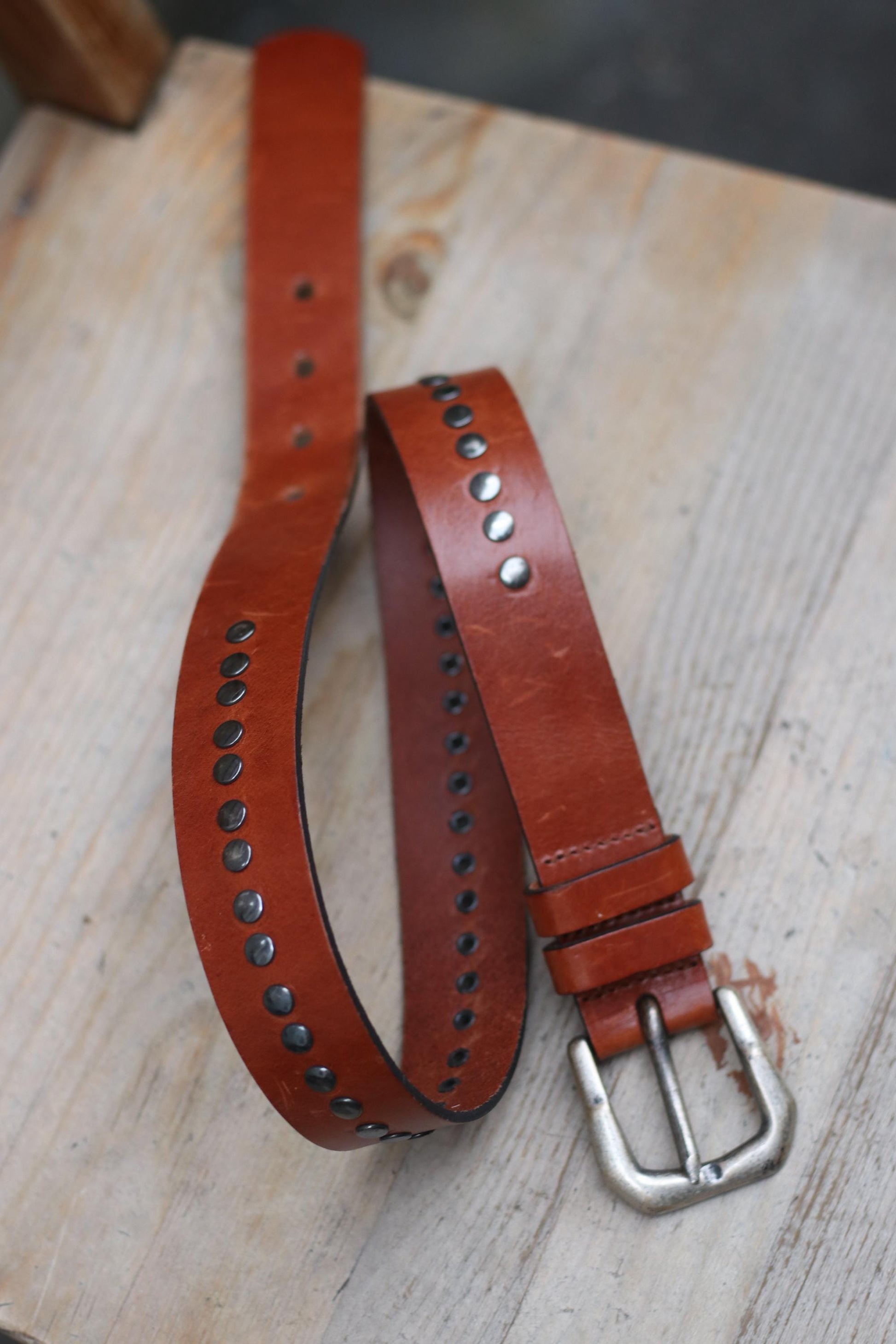 A top view of a vintage handmade brown leather belt with a silver buckle and decorative silver studs is presented on a brown  textured wooden surface.