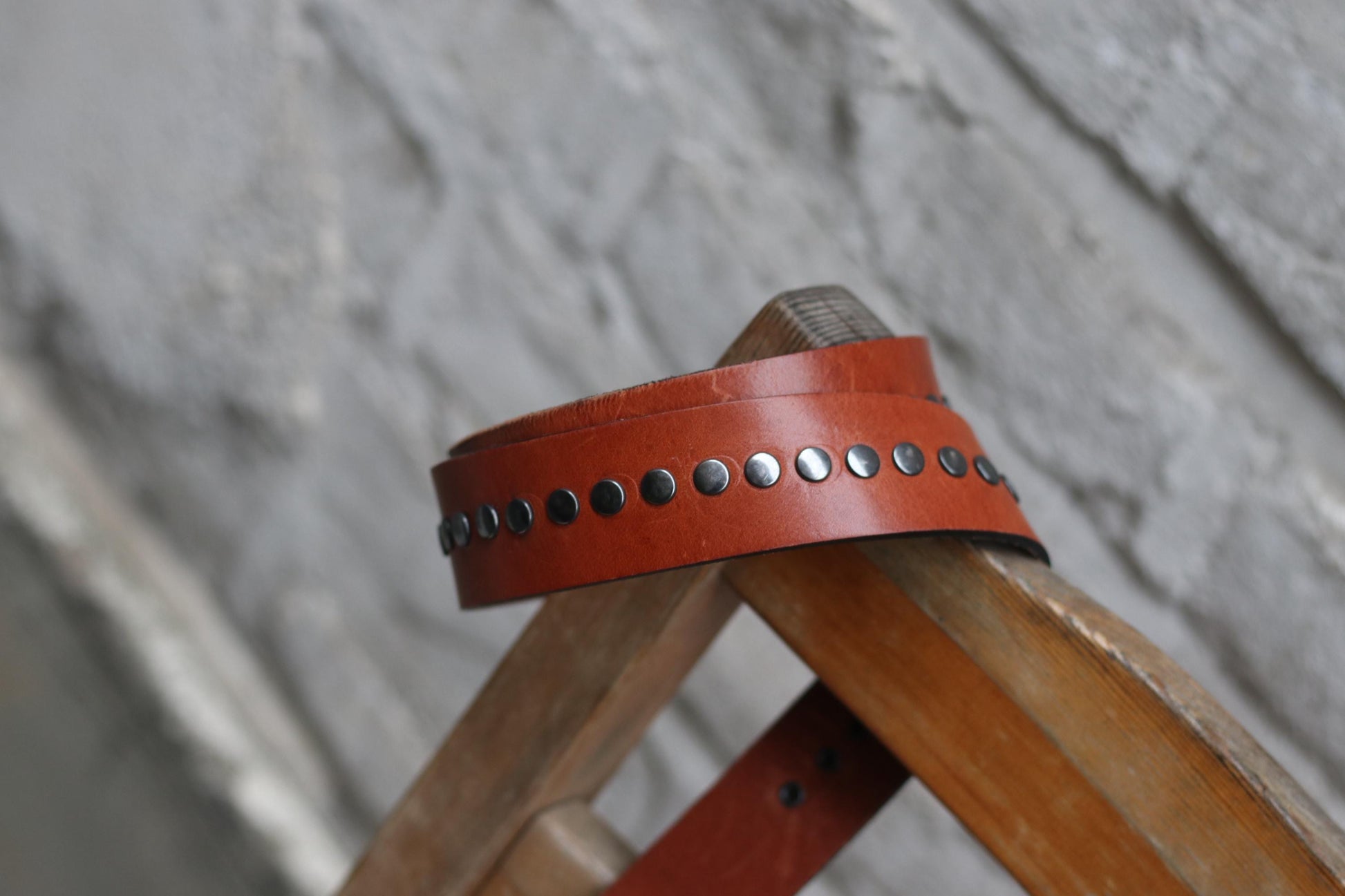 A handmade 70s studded brown leather belt rests on a wooden chair, with a blurred stone wall behind it.