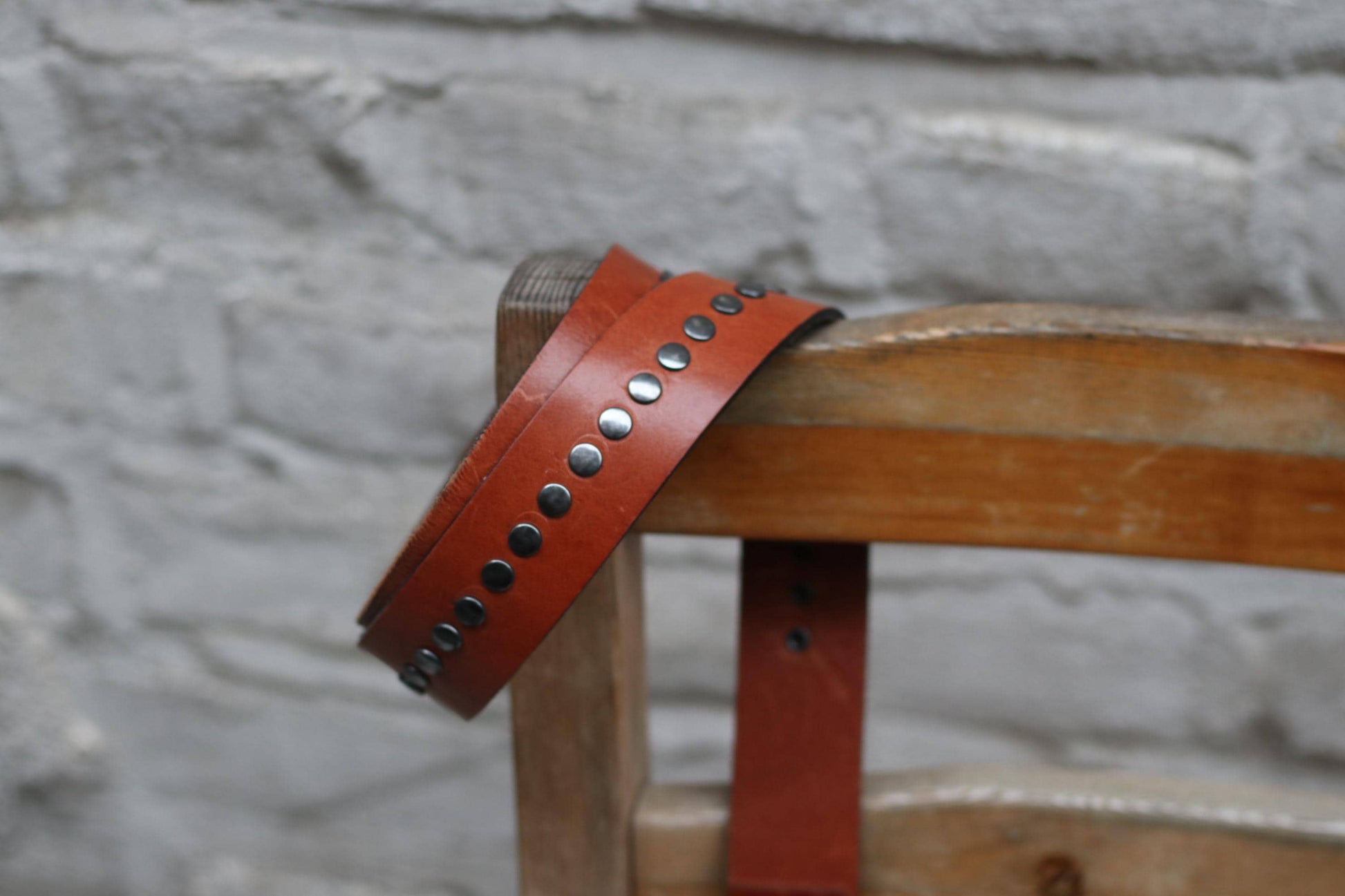 close-up of a brown leather belt with silver studs, draped over the armrest of a wooden chair. A textured stone wall provides a neutral backdrop.