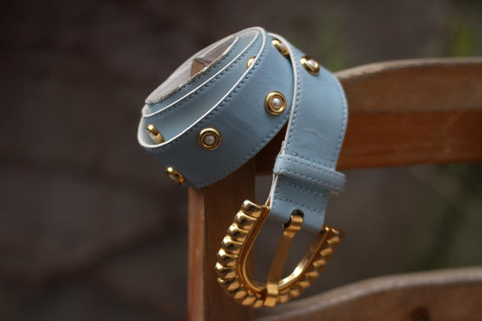A vintage light blue leather Escada belt with a gold-tone scalloped buckle and pearl and gold-tone studs, displayed on a wooden surface.