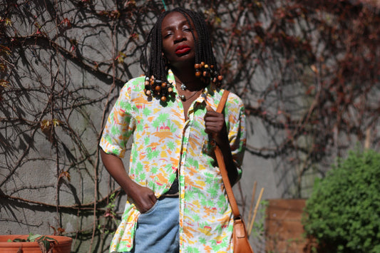 Model with braided hair with beaded accents wearing a yellow and green tropical vintage shirt with denim cut offs and a brown leather bag is photographed against a grey wall with browning ivy