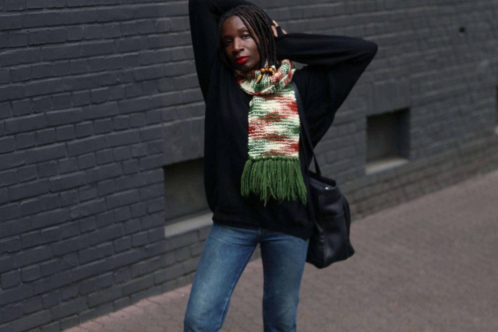 Unisex boho knit scarf with long tassels and a warm, chunky feel, styled  on a female model wearing g jeans and a black sweater, captured against a grey bricked wall