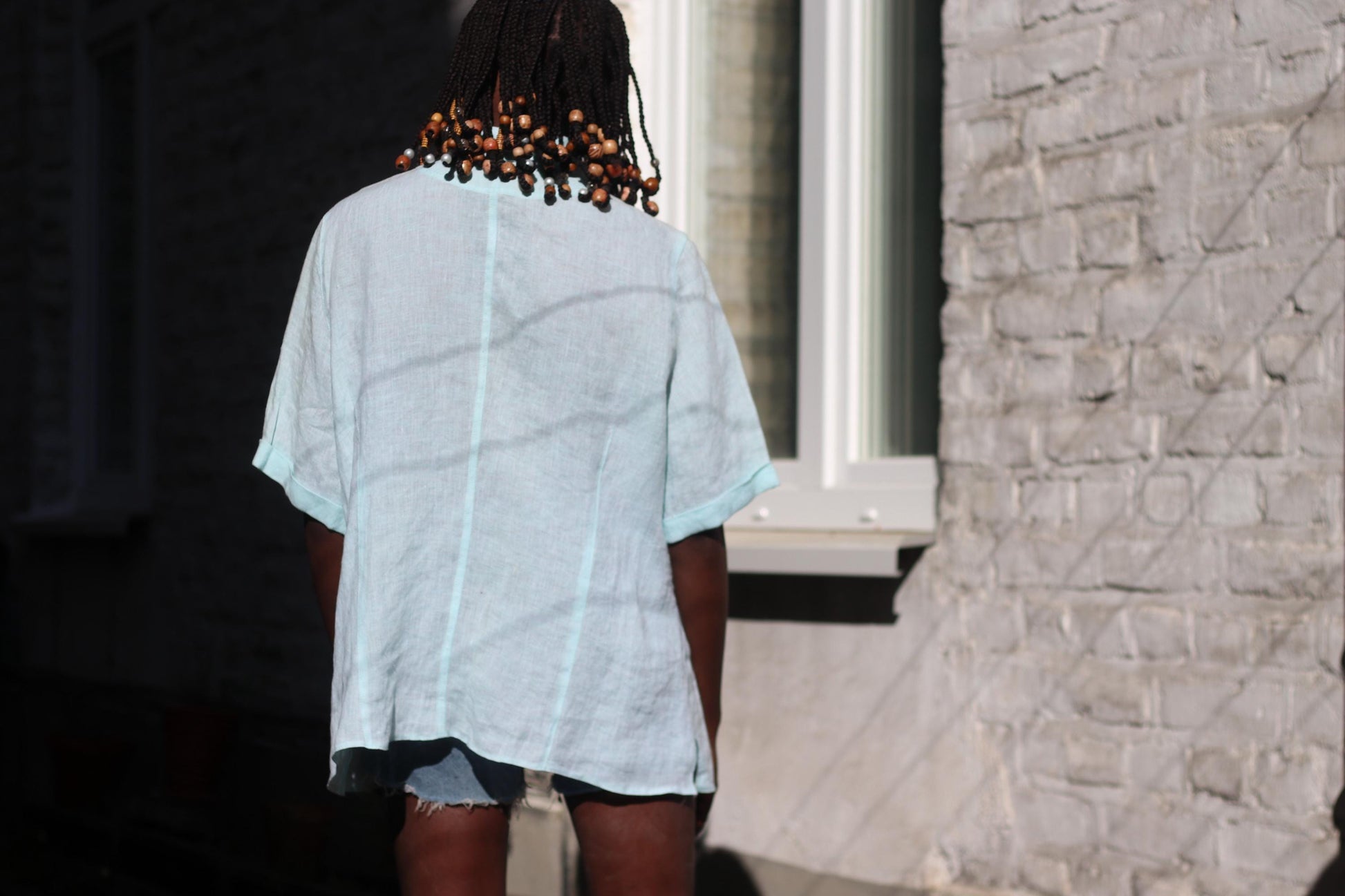 A female model  with braids is photographed from the back in a light blue vintage linen blouse and denim shorts outdoors, with a brick wall and sunlight in the background. The blouse is short-sleeved and the shorts are streetstyle.