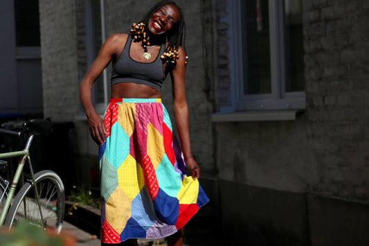 A smiling model of African heritage  with dark braided hair and beads, wearing a gray top cropped and a vintage vibrant, multi-colored handmade patchwork skirt, stands outdoors with hands on the skirt.  The mid-length skirt was made in the 70s