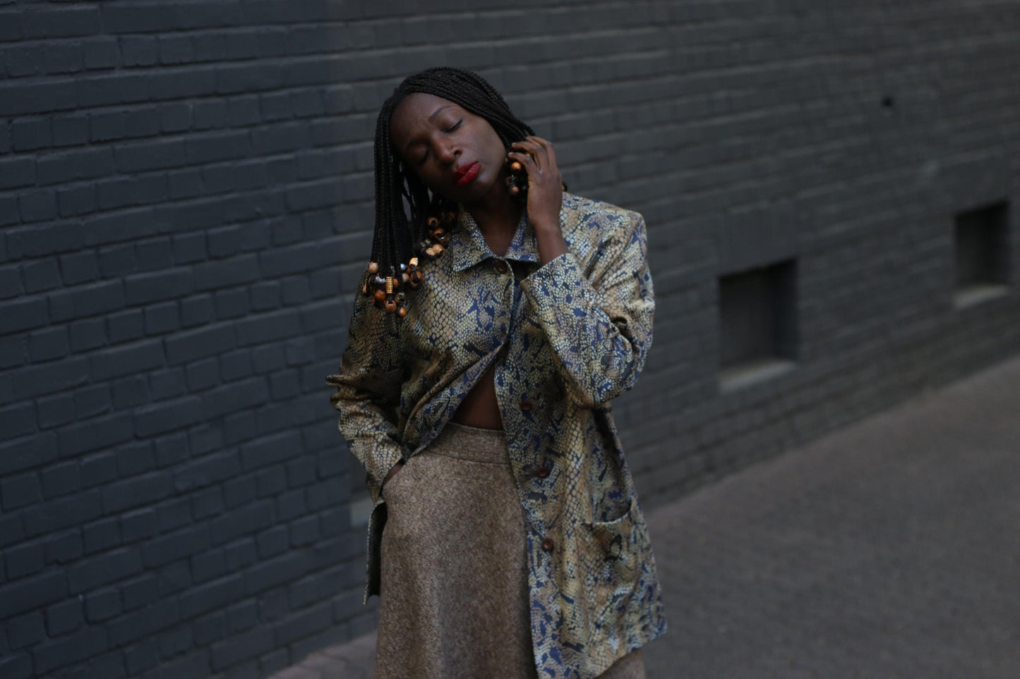 A Female model with beaded braids wearing  a vintage 90s snakeskin duster jacket with a relaxed fit and oversized pockets  with a brow  skirt photographed against  a grey brick wall to showcase the bold yet neutral layering piece perfect for standout street style