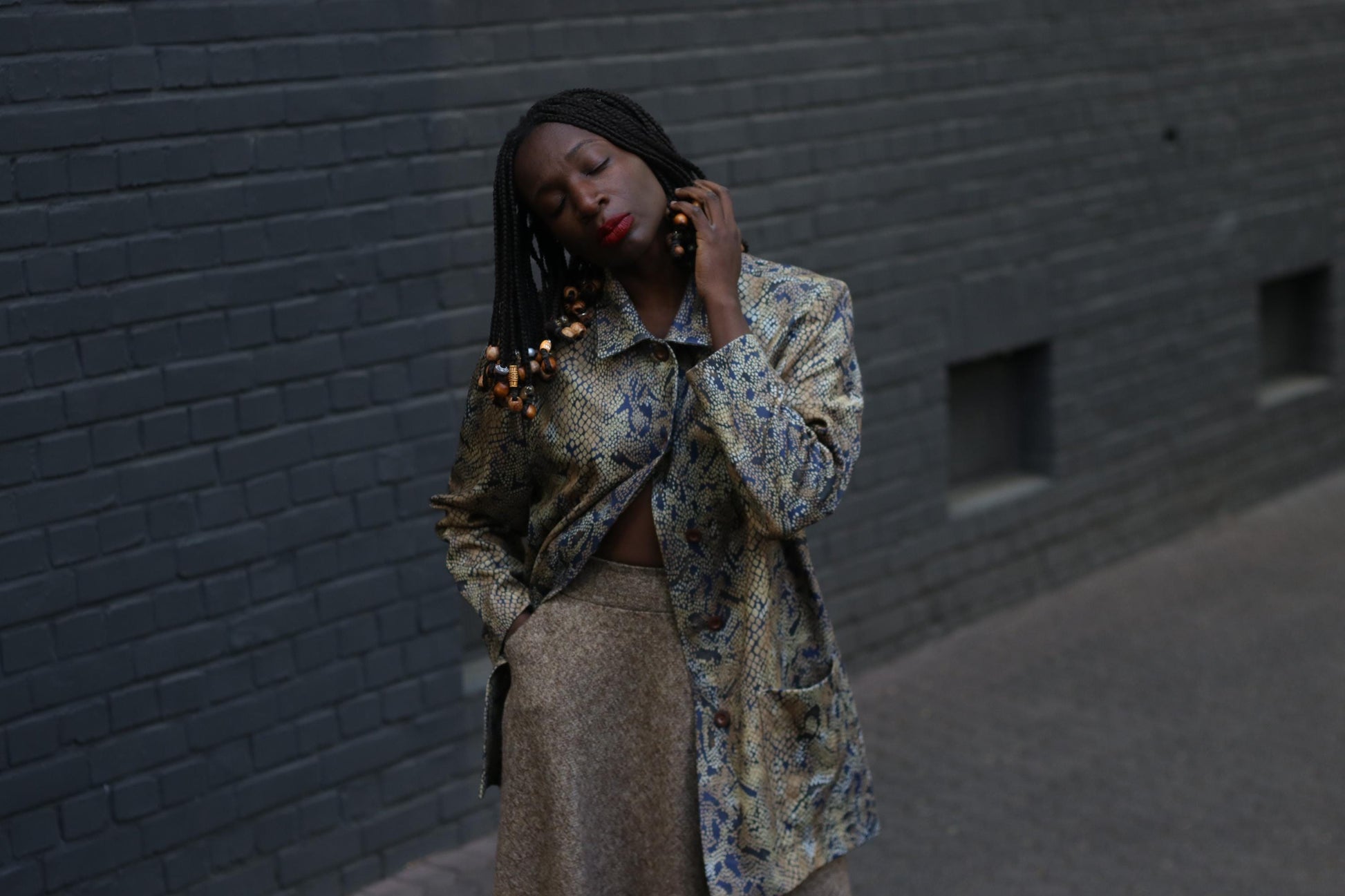 A Female model with beaded braids wearing  a vintage 90s snakeskin duster jacket with a relaxed fit and oversized pockets  with a brow  skirt photographed against  a grey brick wall to showcase the bold yet neutral layering piece perfect for standout street style
