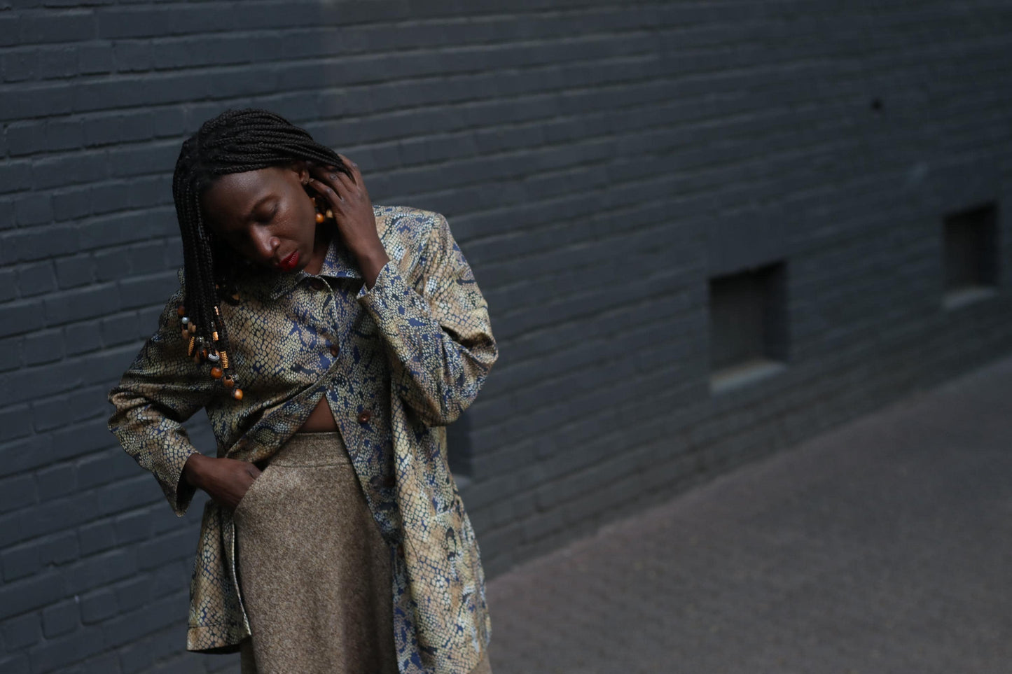 Female model with beaded braids  in a vintage 90s snakeskin duster jacket with a relaxed fit and oversized pockets photographed against  a grey brick wall to showcase the bold yet neutral layering piece perfect for standout street style