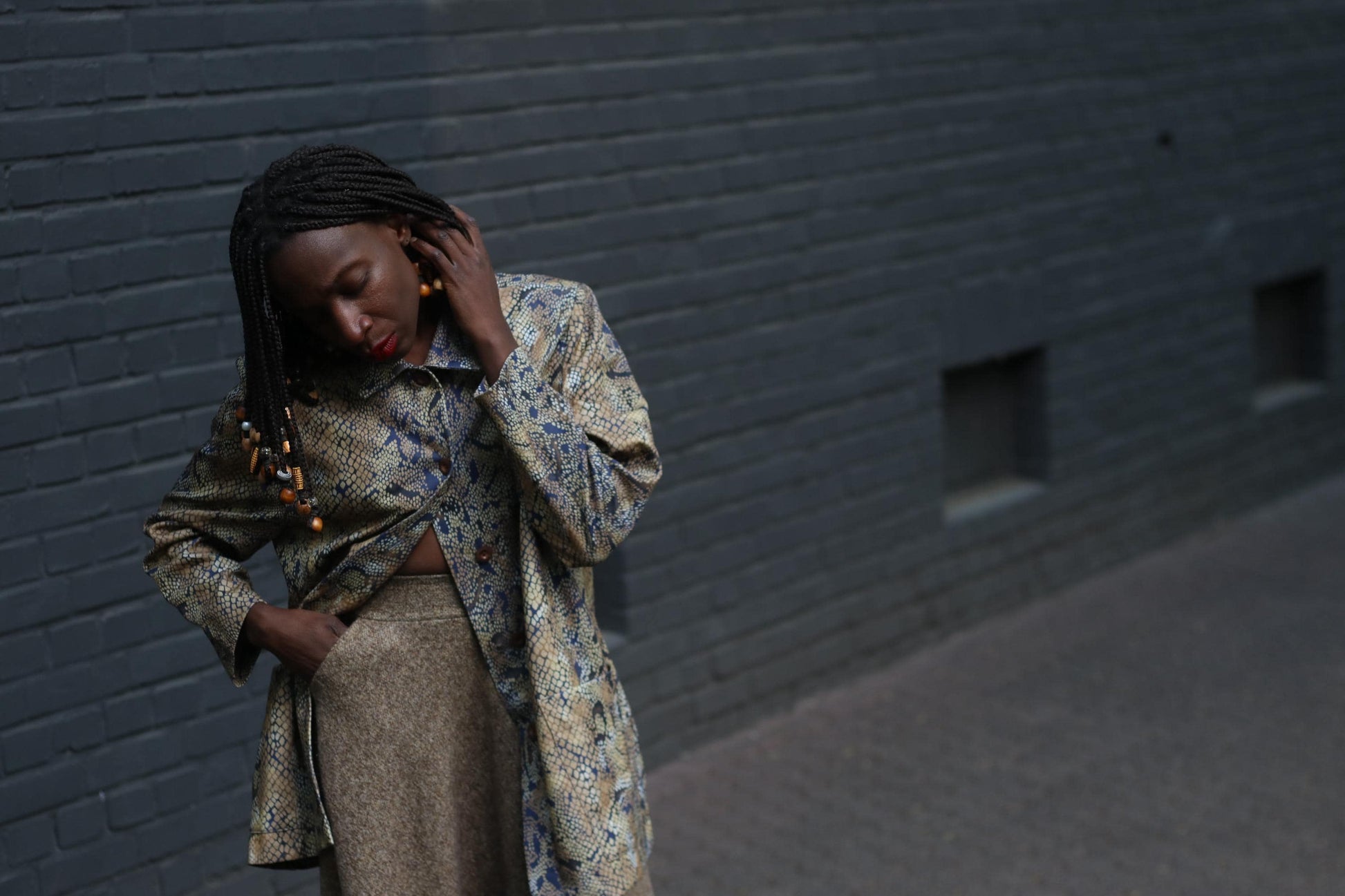 Female model with beaded braids  in a vintage 90s snakeskin duster jacket with a relaxed fit and oversized pockets photographed against  a grey brick wall to showcase the bold yet neutral layering piece perfect for standout street style