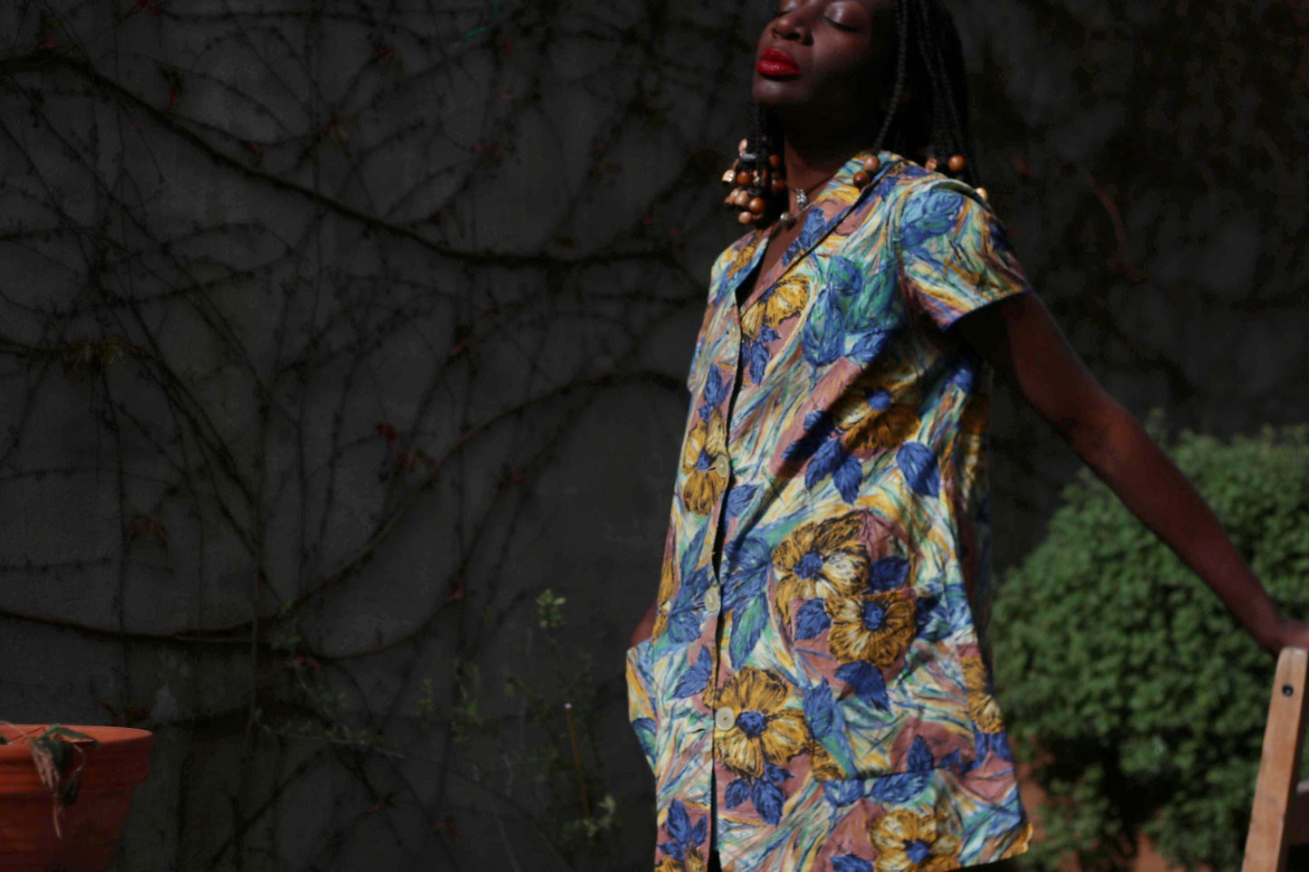 Woman in a vintage floral print tunic dress with a blue and yellow palette, captured outdoors in a garden against a wall with browning twines, glowing in soft light with a nostalgic 70s feel.