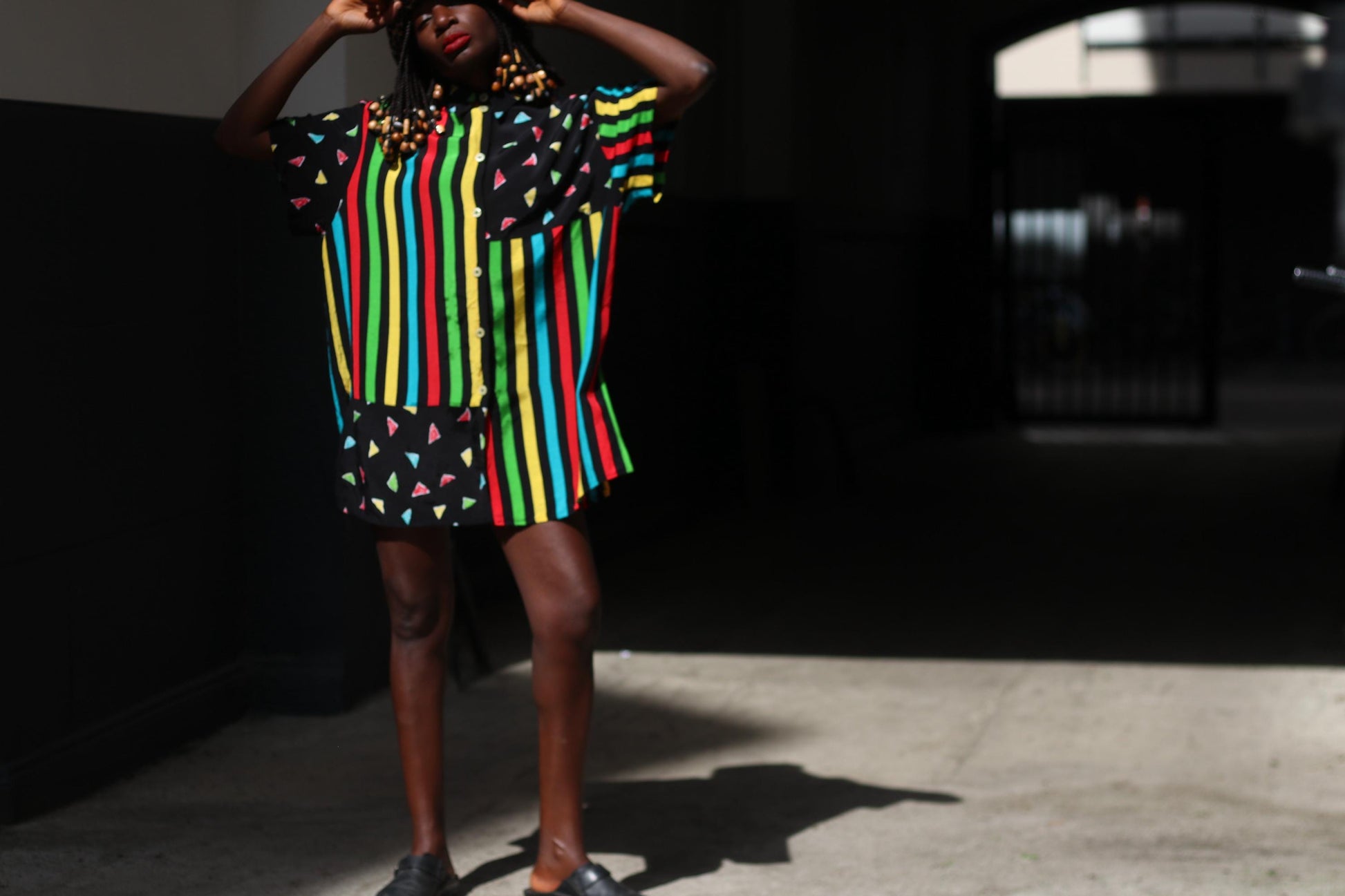 A female model with dark skin and red lipstick models a colorful 90s oversized shirt that can be worn as a dress. The retro button-up has vertical stripes of red, yellow, green, and blue and features black panels with colorful triangles. The model accessories the look with an animal print bucket hat. The image is shot in a sunlight alley