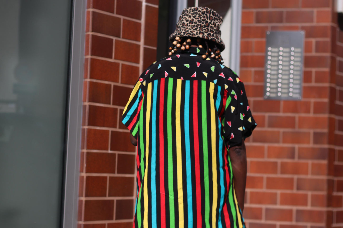 A back view of a  female model in a colorful 90s oversized shirt that can be worn as a dress. The retro button-up has  vertical stripes of red, yellow, green, and blue and features black panels with colorful triangles. The model accessories the look with an animal print bucket hat. The image is shot outdoors against brown tiled walls