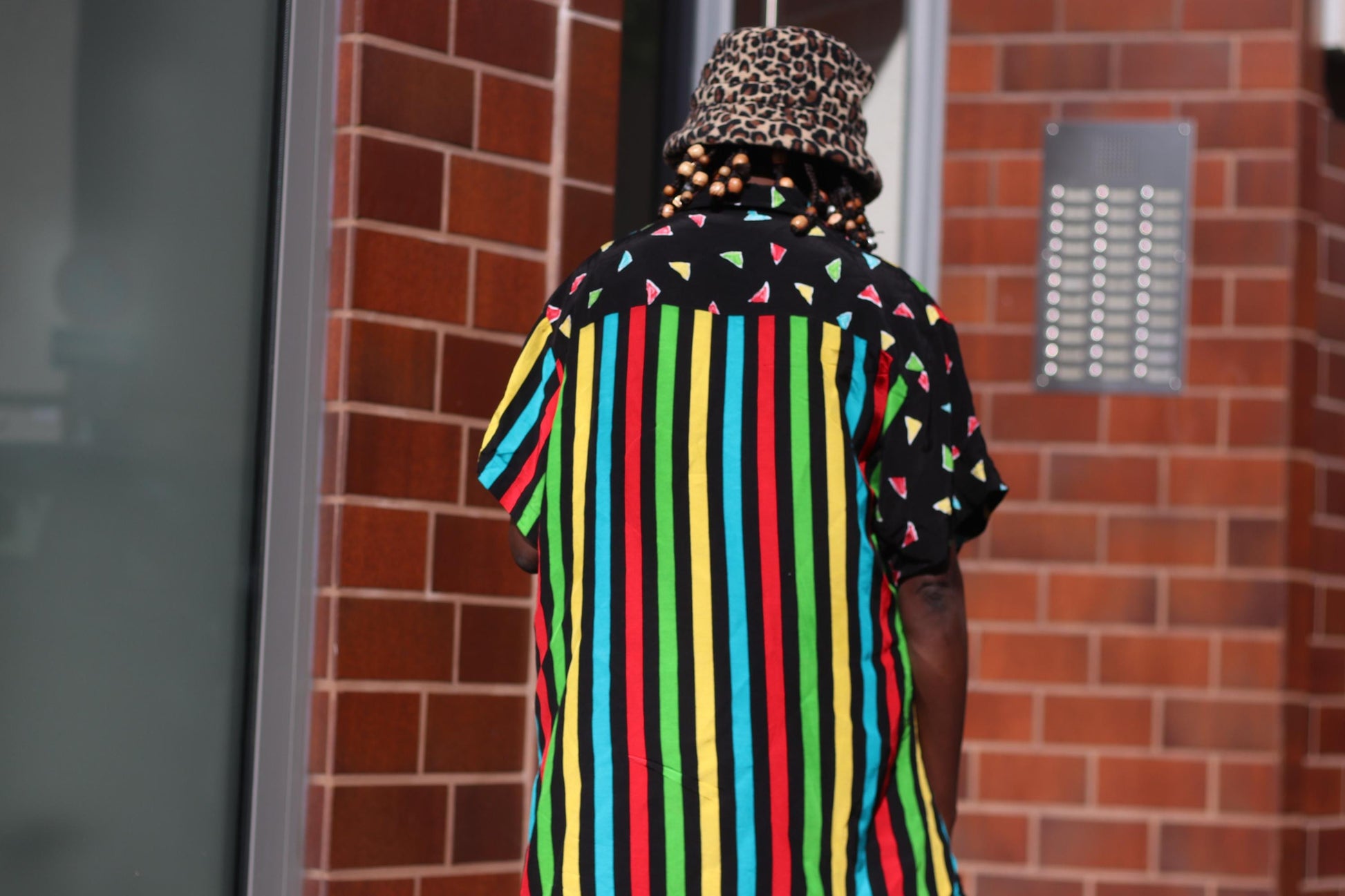 A back view of a  female model in a colorful 90s oversized shirt that can be worn as a dress. The retro button-up has  vertical stripes of red, yellow, green, and blue and features black panels with colorful triangles. The model accessories the look with an animal print bucket hat. The image is shot outdoors against brown tiled walls
