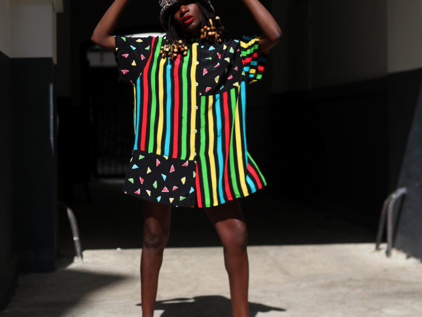 A female model with dark skin and red lipstick models a colorful 90s oversized shirt that can be worn as a dress. The retro button-up has  vertical stripes of red, yellow, green, and blue and features black panels with colorful triangles. The model accessories the look with an animal print bucket hat. The image is shot in a sunlight alley