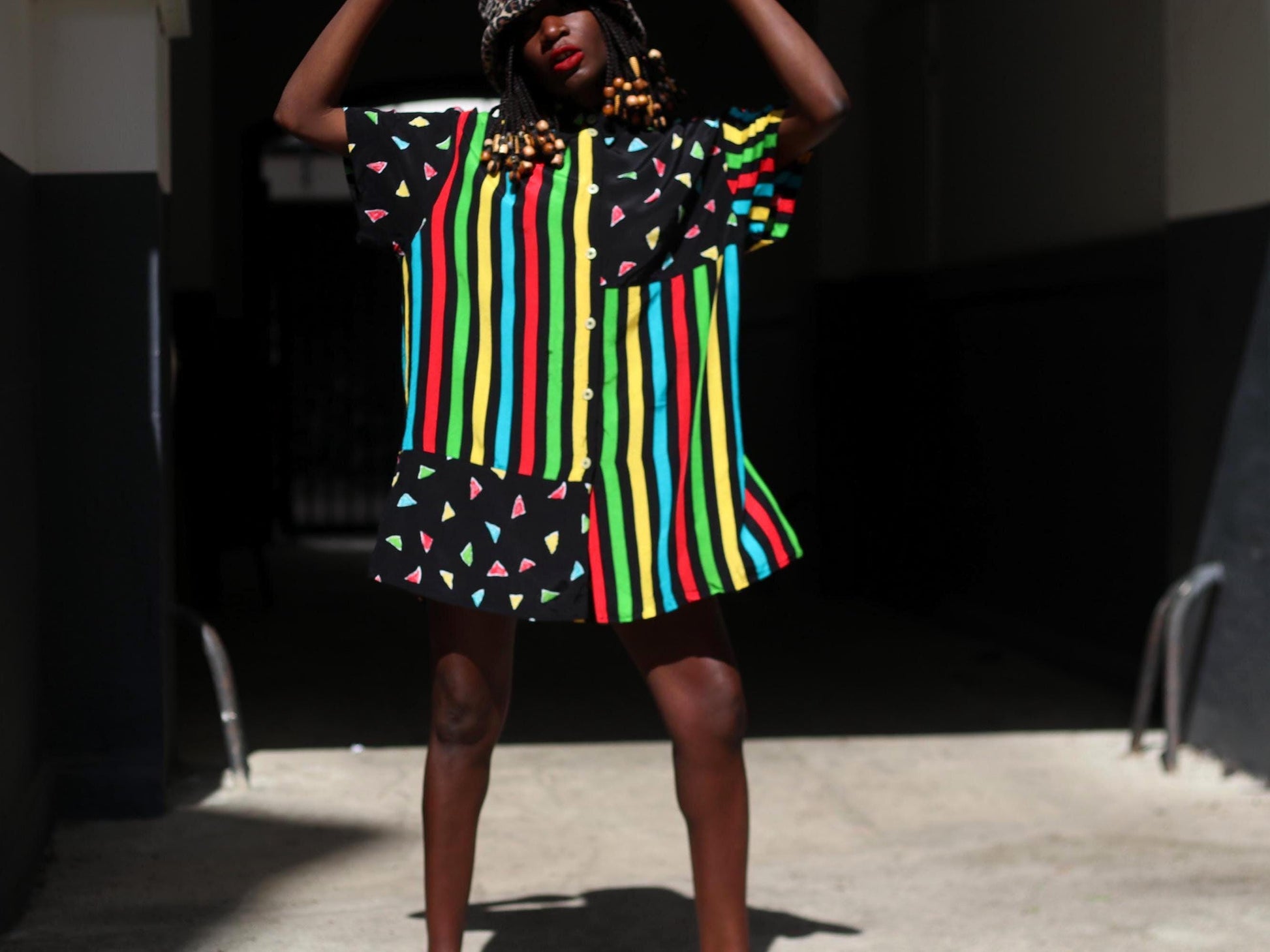A female model with dark skin and red lipstick models a colorful 90s oversized shirt that can be worn as a dress. The retro button-up has  vertical stripes of red, yellow, green, and blue and features black panels with colorful triangles. The model accessories the look with an animal print bucket hat. The image is shot in a sunlight alley
