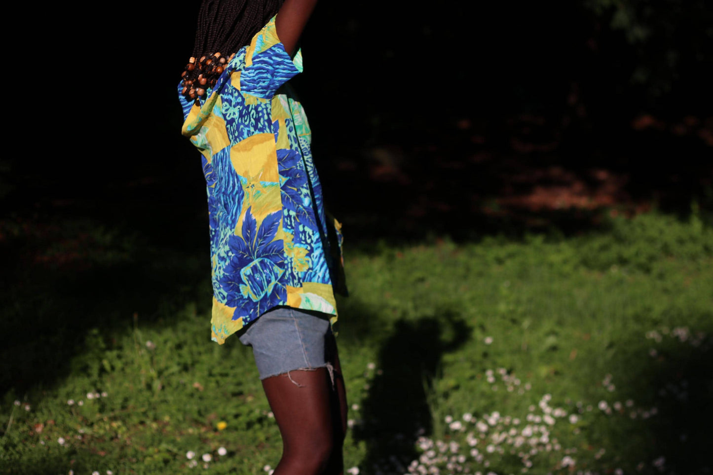 Retro 90s colorful tropical blouse with short sleeves and patchwork design, modeled in sunny garden setting. The image is a side view of the model who styled the shirt with blue denim shorts.