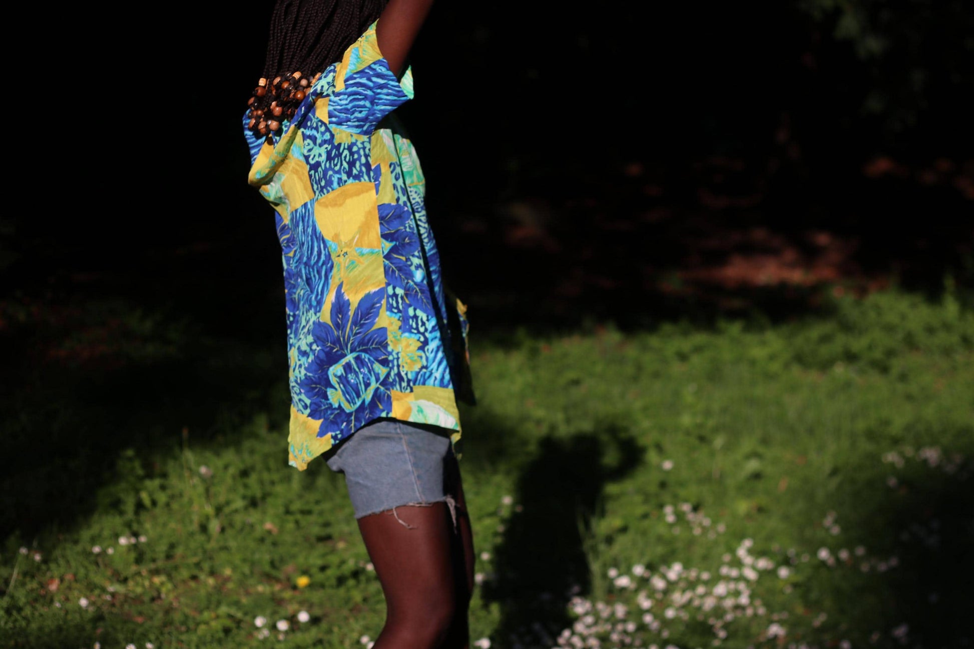 Retro 90s colorful tropical blouse with short sleeves and patchwork design, modeled in sunny garden setting. The image is a side view of the model who styled the shirt with blue denim shorts.