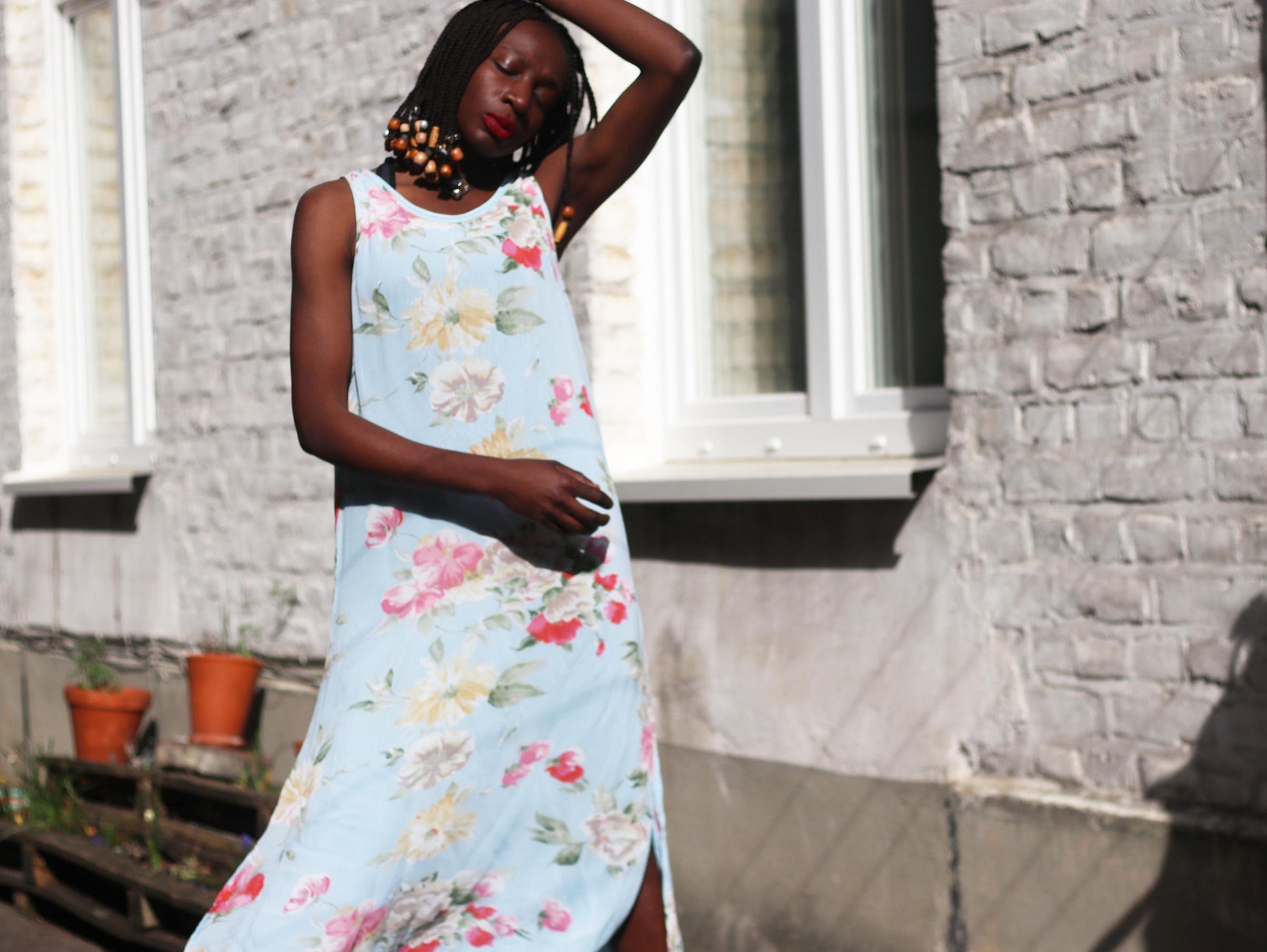 Female model of african heritage with braided hair wearing a 90s Cottagecore maxi dress with a sky blue floral pattern. The summer dress has a layered hem and is sleeveless. The woman is outdoors and photographed against a brick wall and windows with a dreamy expression
