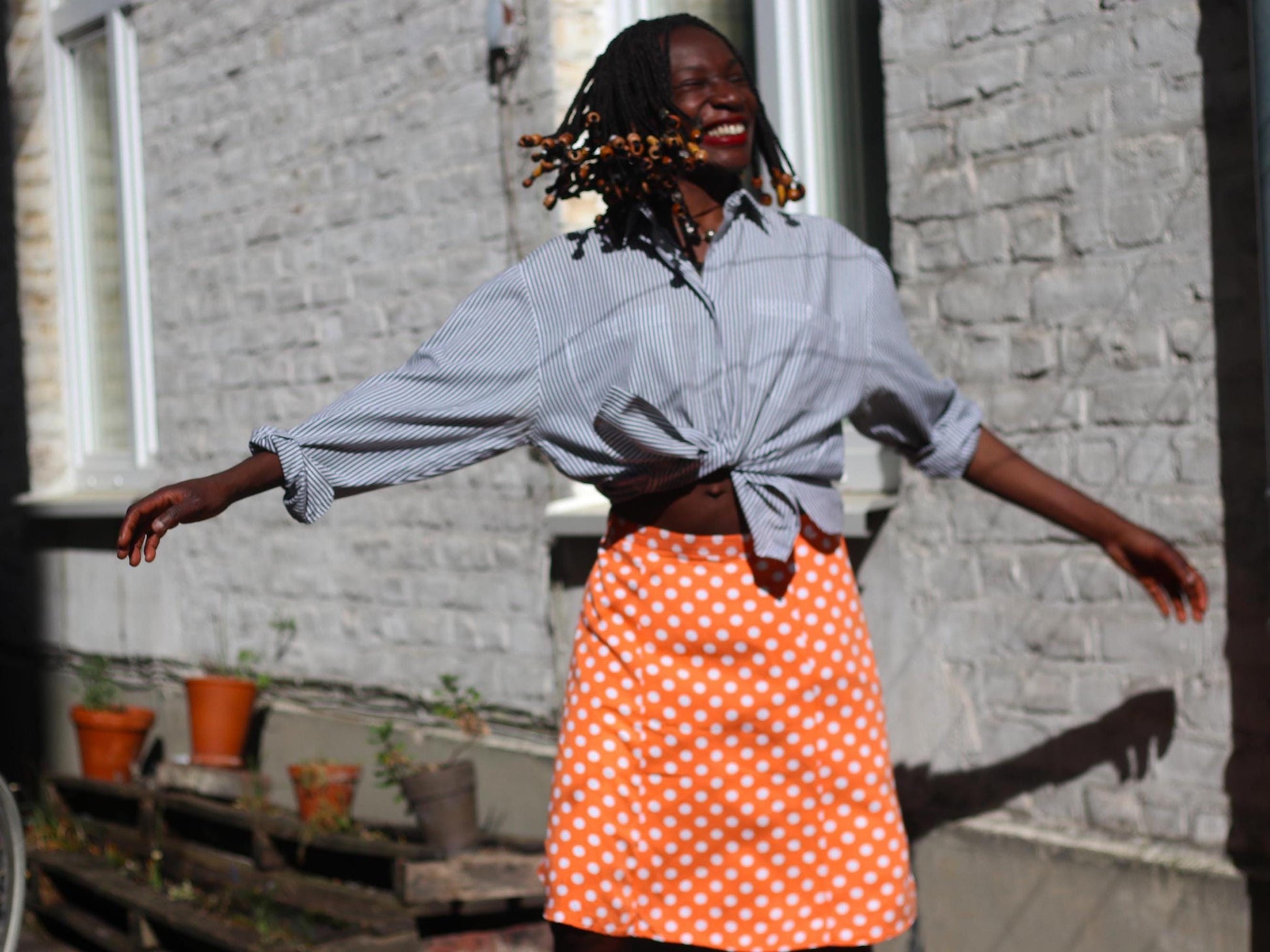 A female model wearing a vintage orange polka dot skirt and a 90s pinstripe blouse, photographed outdoors. She is joyful with her hands out like she is about to spin. The skirt is knee-length and flirty and fun exuding a french chic vibe